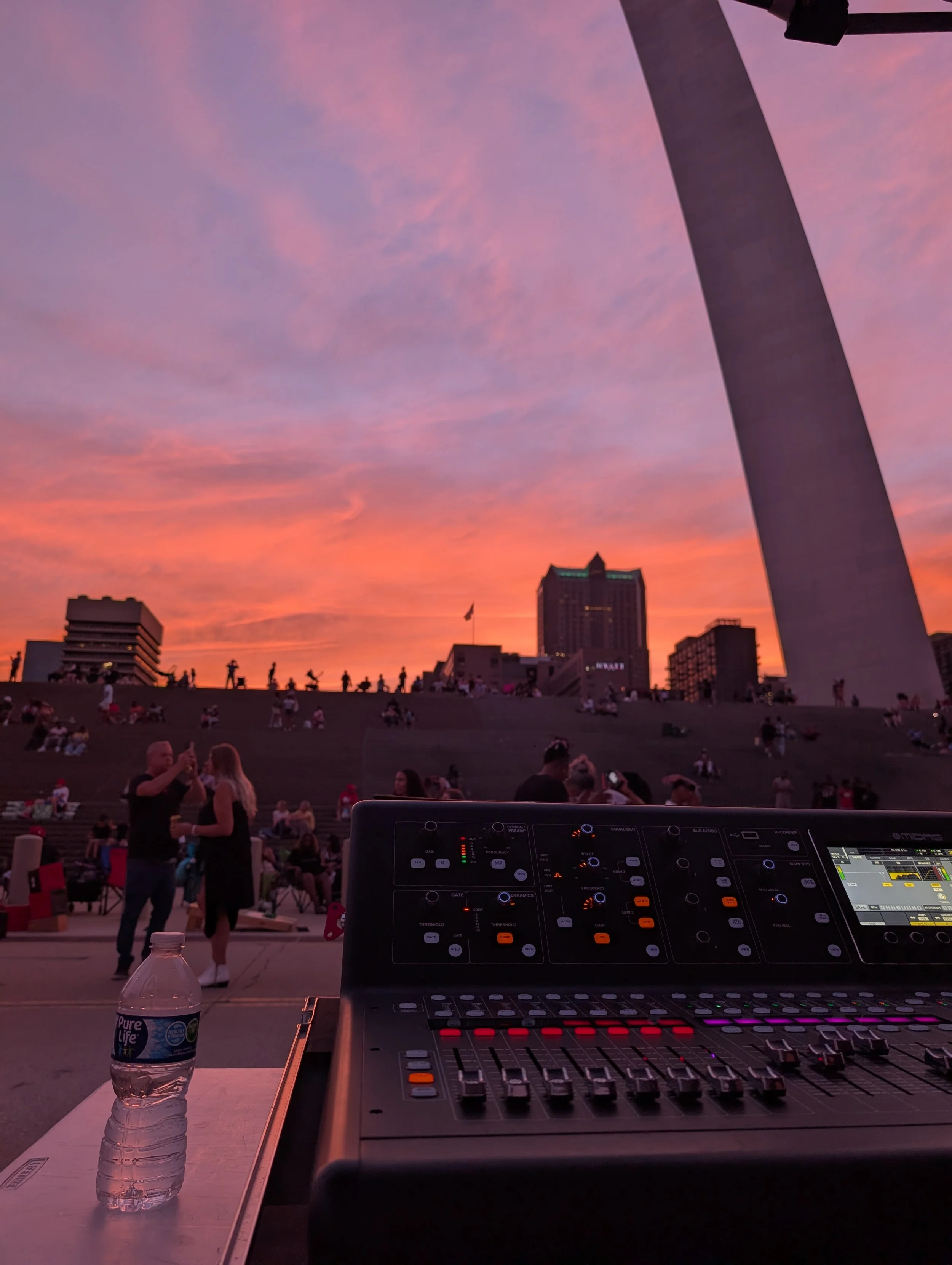 A digital music mixer on a table at an outdoor concert with a city skyline and sunset in the background. silhouettes of people are visible on a terrace, and a water bottle is in the foreground.