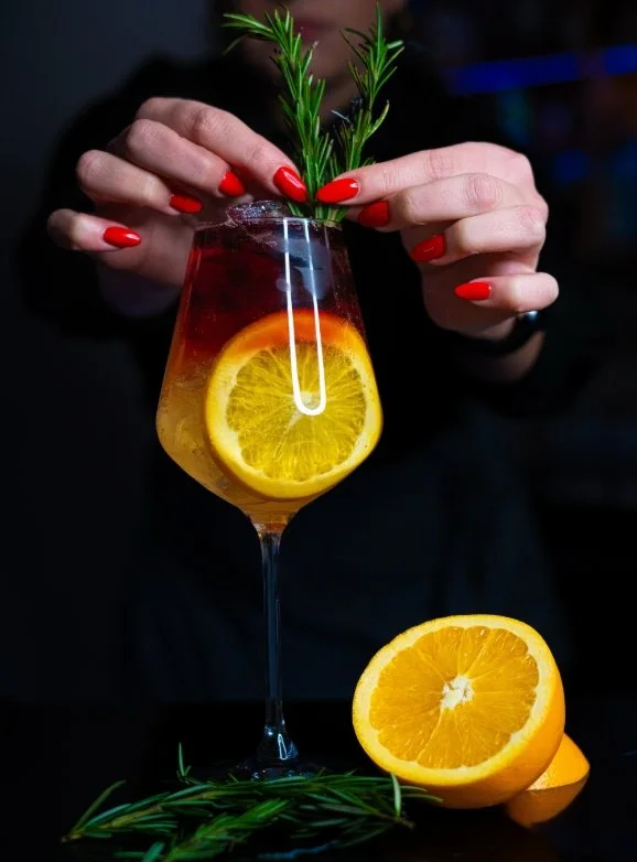 A person with red painted nails holds a cocktail glass garnished with orange slices and a sprig of rosemary.