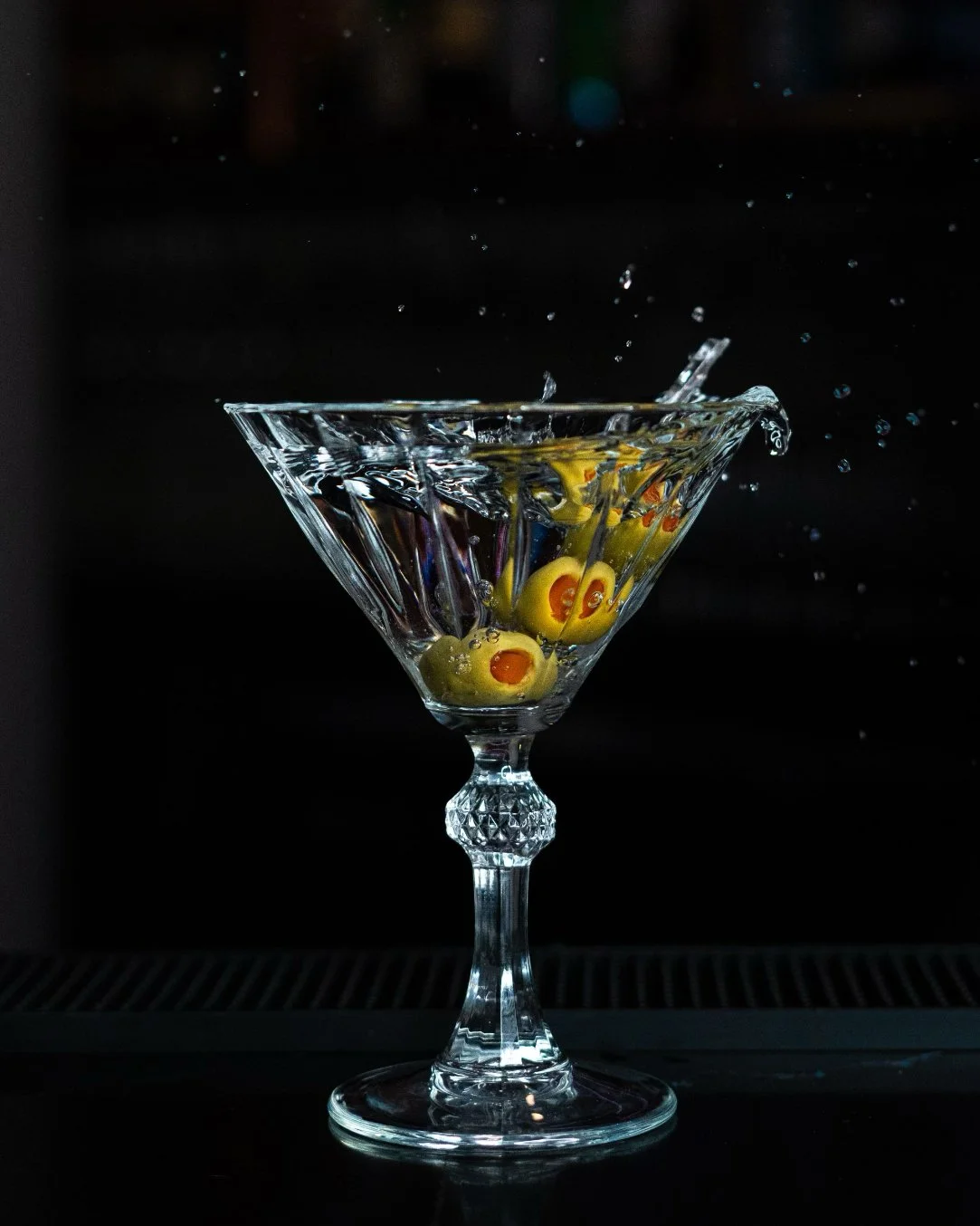 A crystal cocktail with floating olives, and water splashing outside the glass against a black background.