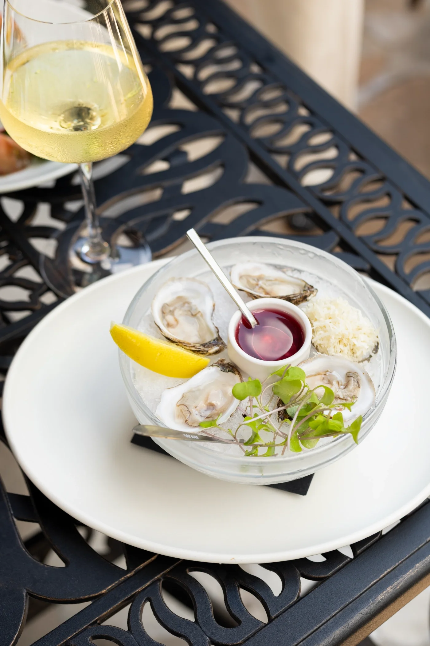A glass of white wine and a plate of oysters on a white plate on a black outdoor table.