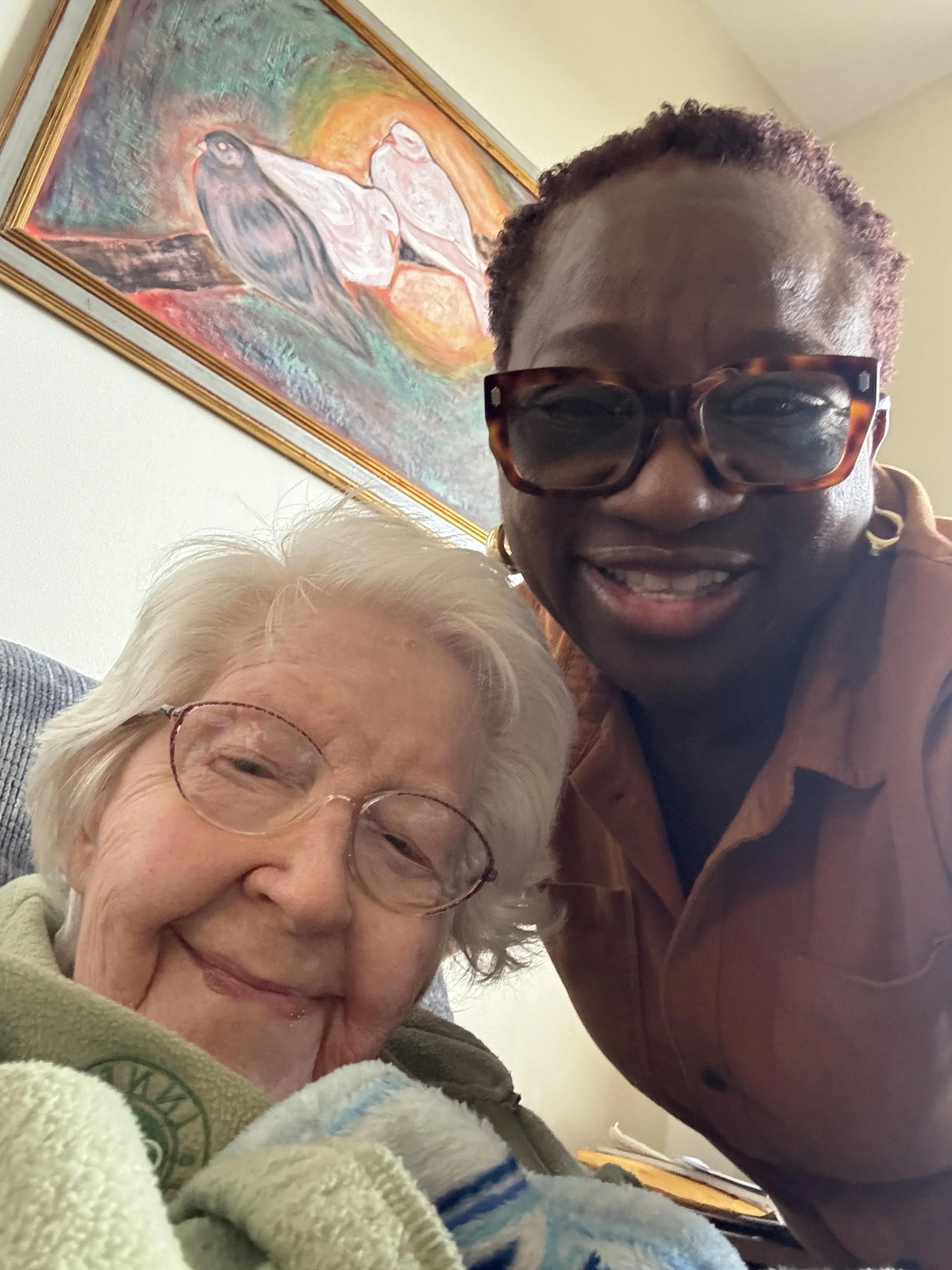 A smiling elderly woman with glasses and white hair, and a smiling African American woman with glasses and short curly hair, taking a selfie together indoors, with a colorful abstract painting hanging on the wall behind them.
