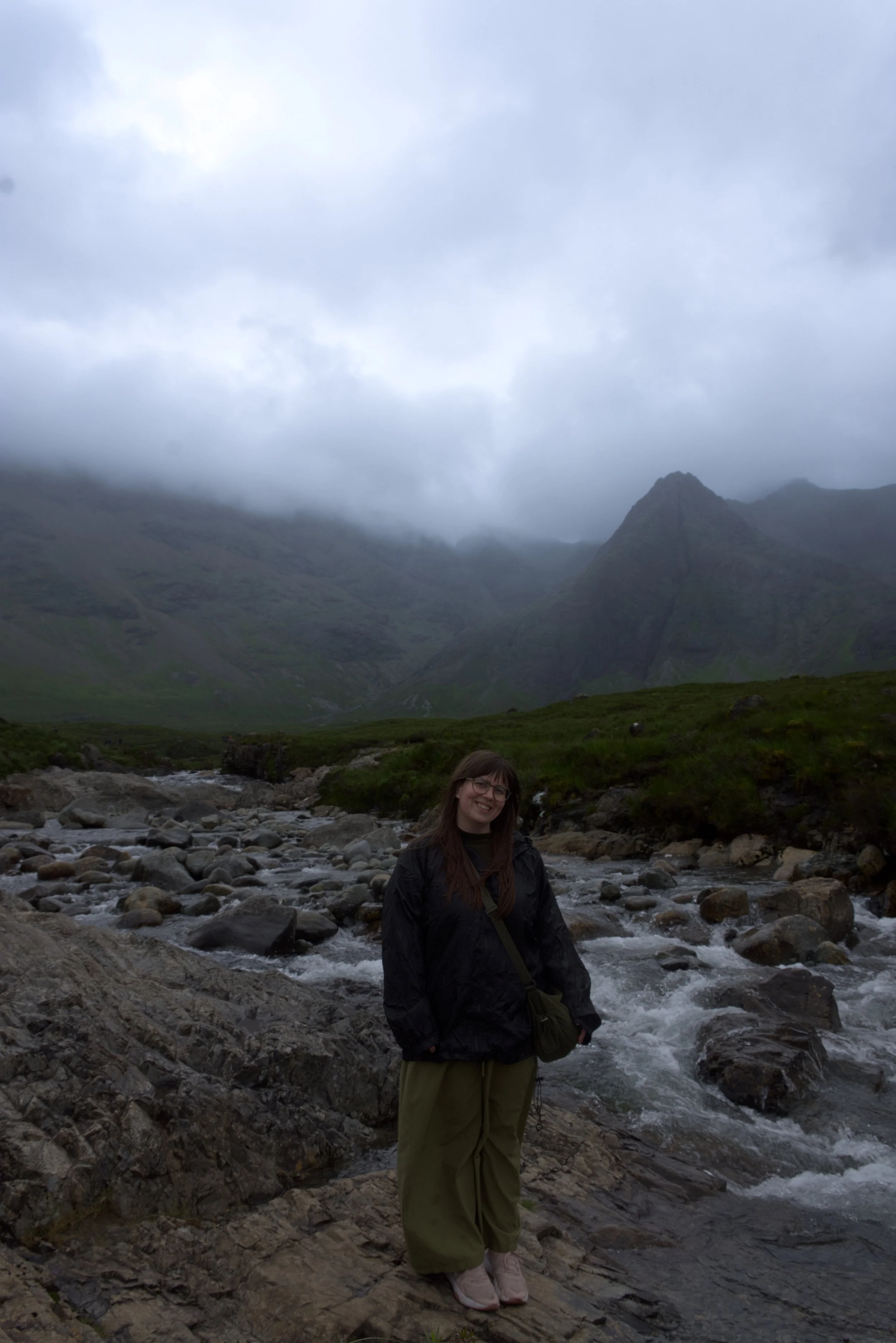 Charlene, freelancer and face of Charlene Shillan Marketing, the photo shoes her with glasses and brown hair standing on rocky terrain by a flowing mountain stream, with foggy mountains and cloudy sky in the background - Skye's Fairypools.