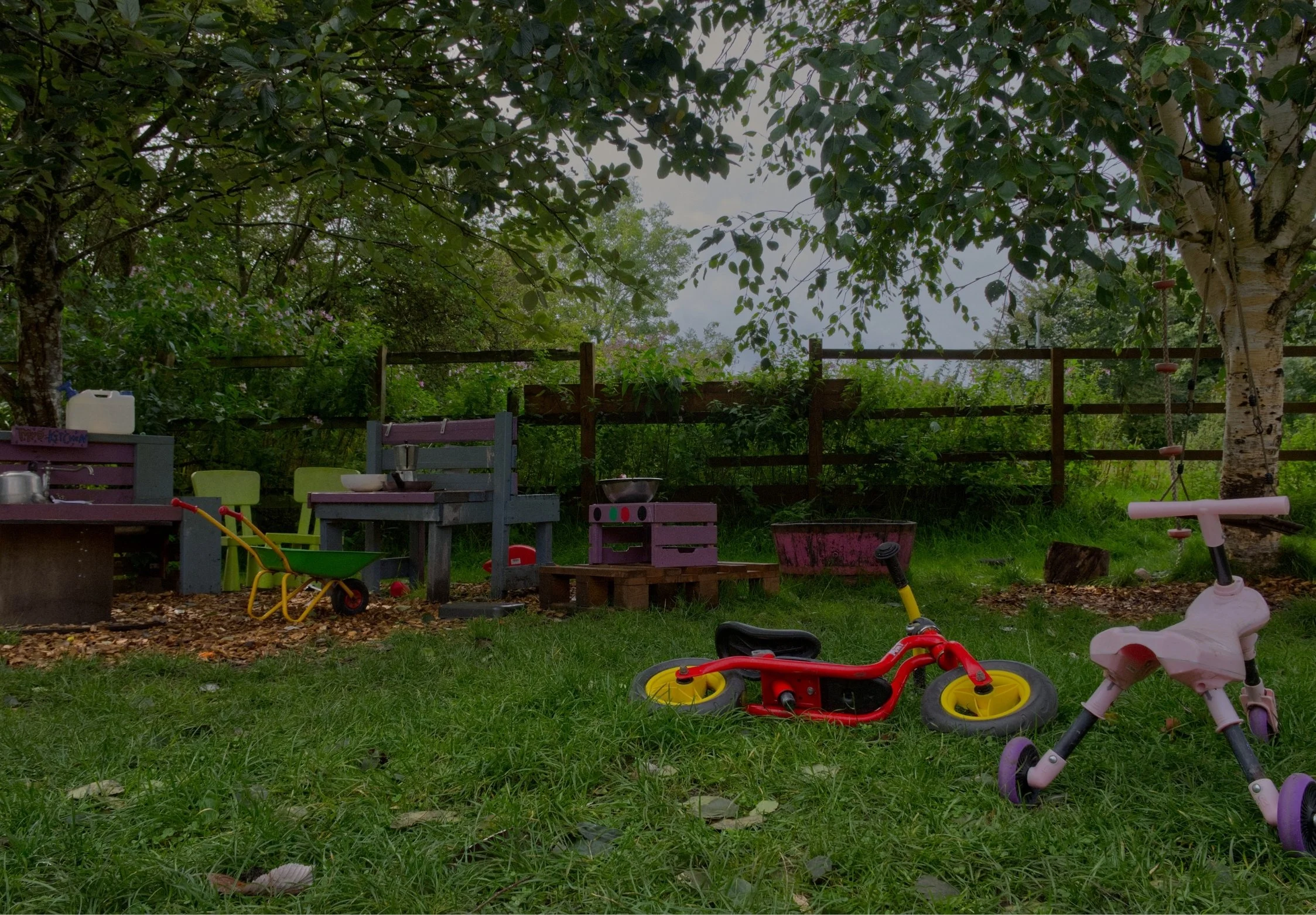 Child's outdoor play area with scattered toys, picnic table, and lush green trees in the background.