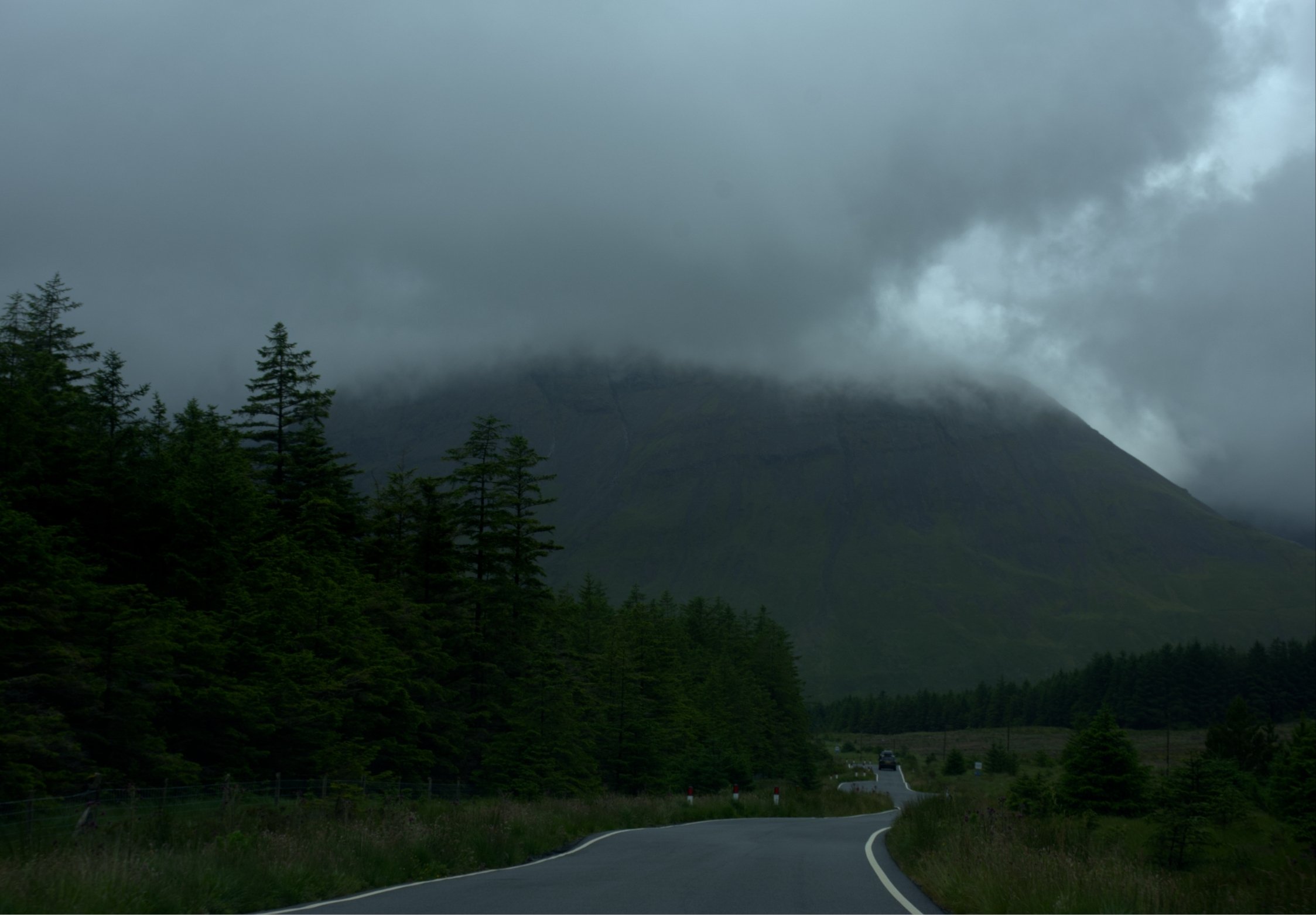 A winding mountain road on the Isle of Skye with a guardrail, surrounded by dense green trees, and a dark cloudy sky with mountains partially obscured by mist or fog.