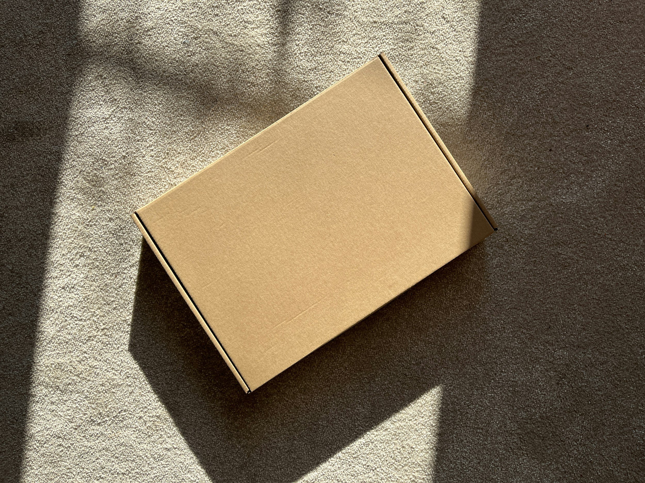 A plain brown cardboard box on a gray carpeted floor with sunlight and shadows casting across it.