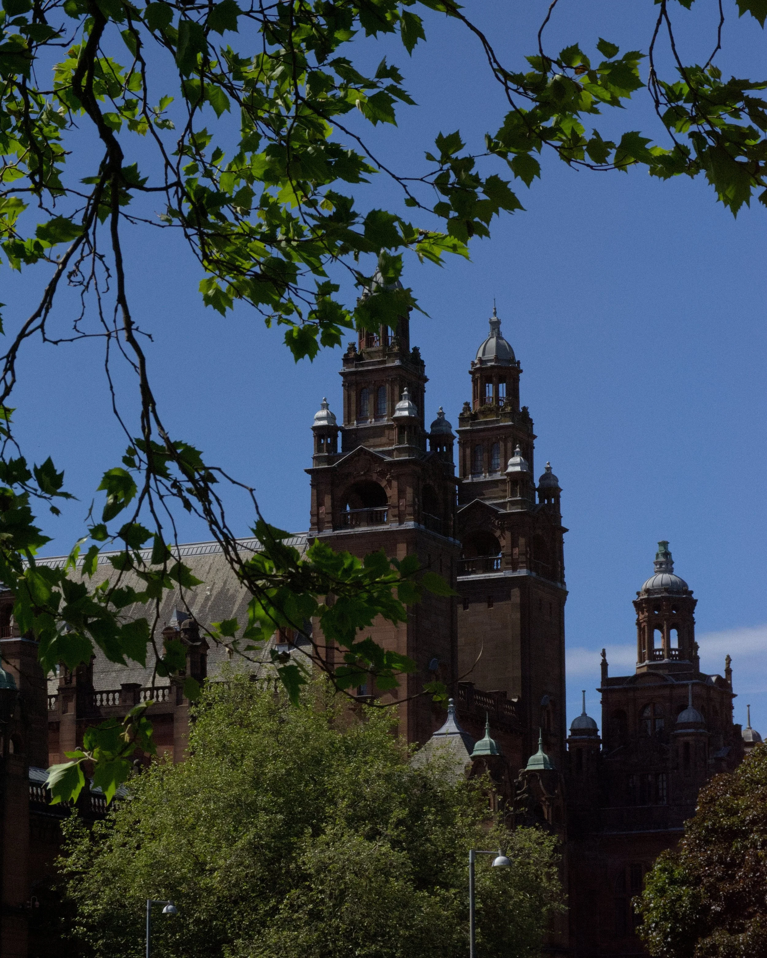 Kelvingrove Art Museum, historic building with multiple towers and ornate architecture, partially obscured by green tree branches and leaves under a clear blue sky.