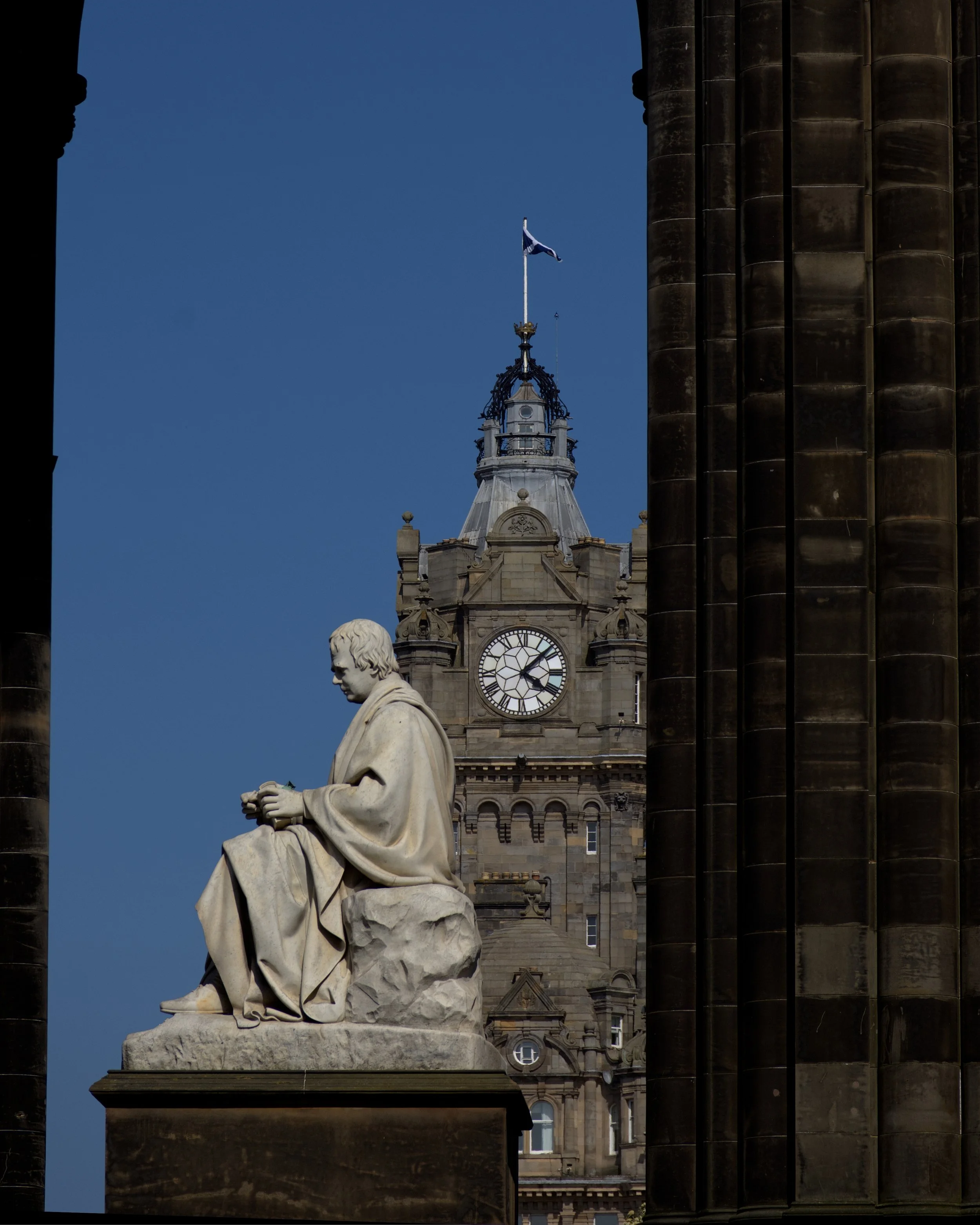 Scott Monument in Edinburgh, a white marble seated statue of a man in front of a historic building with a clock tower, flanked by dark stone columns and archways, with blue sky in the background.