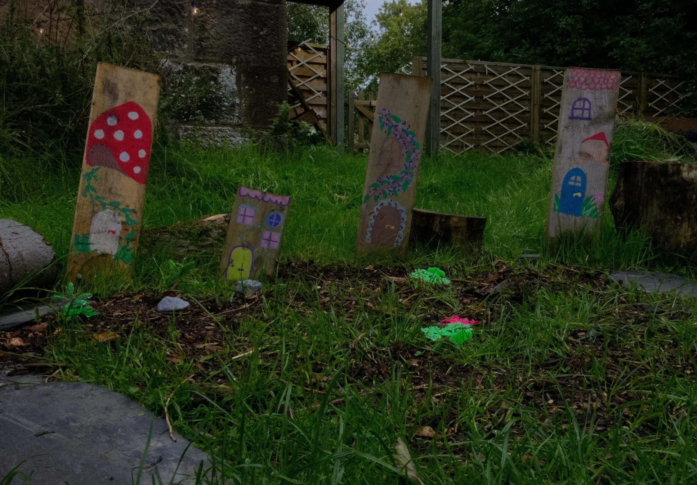 Outdoor garden scene at dusk with painted wooden stakes featuring colorful house and mushroom designs, surrounded by grass, rocks, and trees.