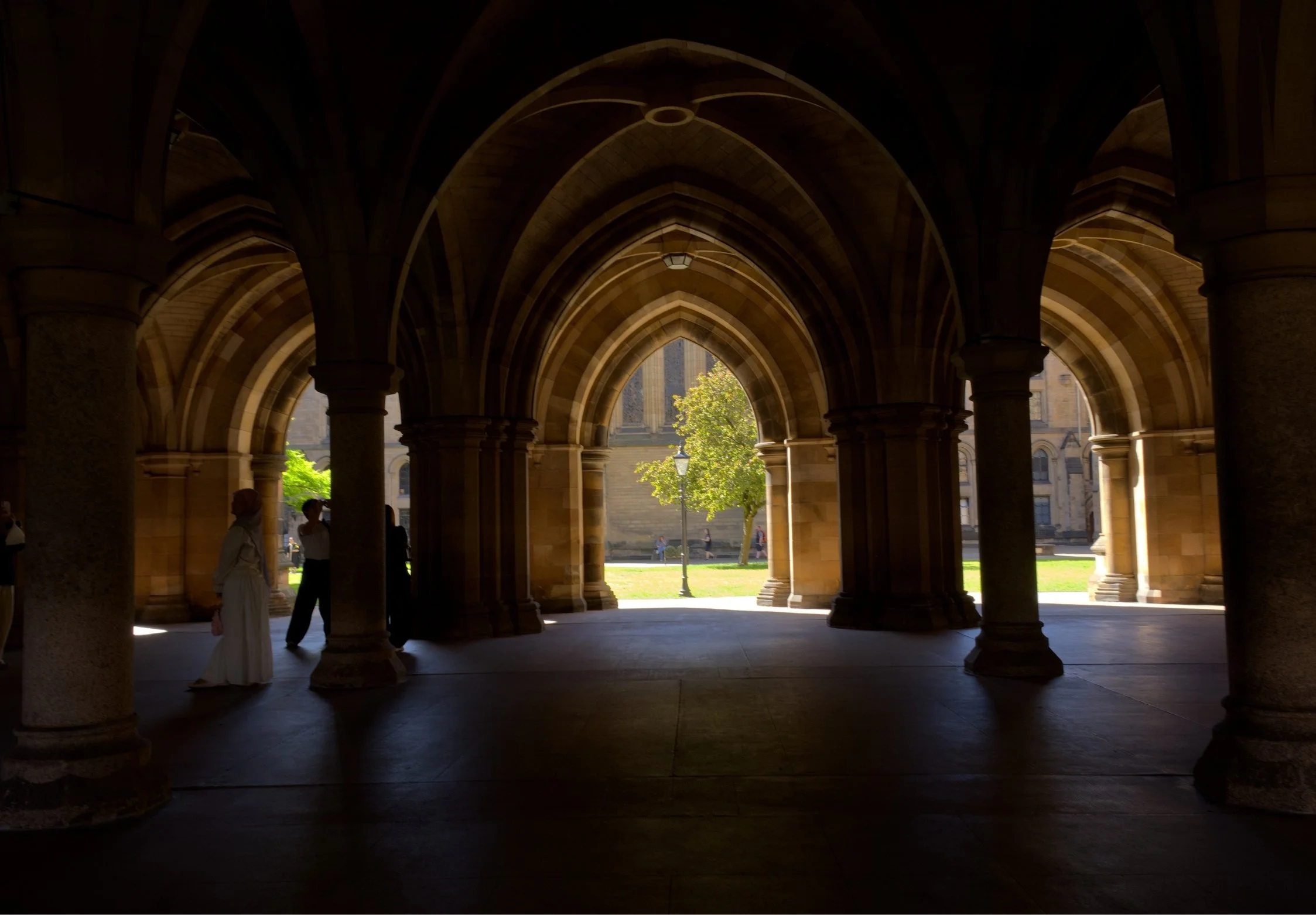View through stone arches of Glasgow University courtyard with people walking and a tree, with a historic building in the background.