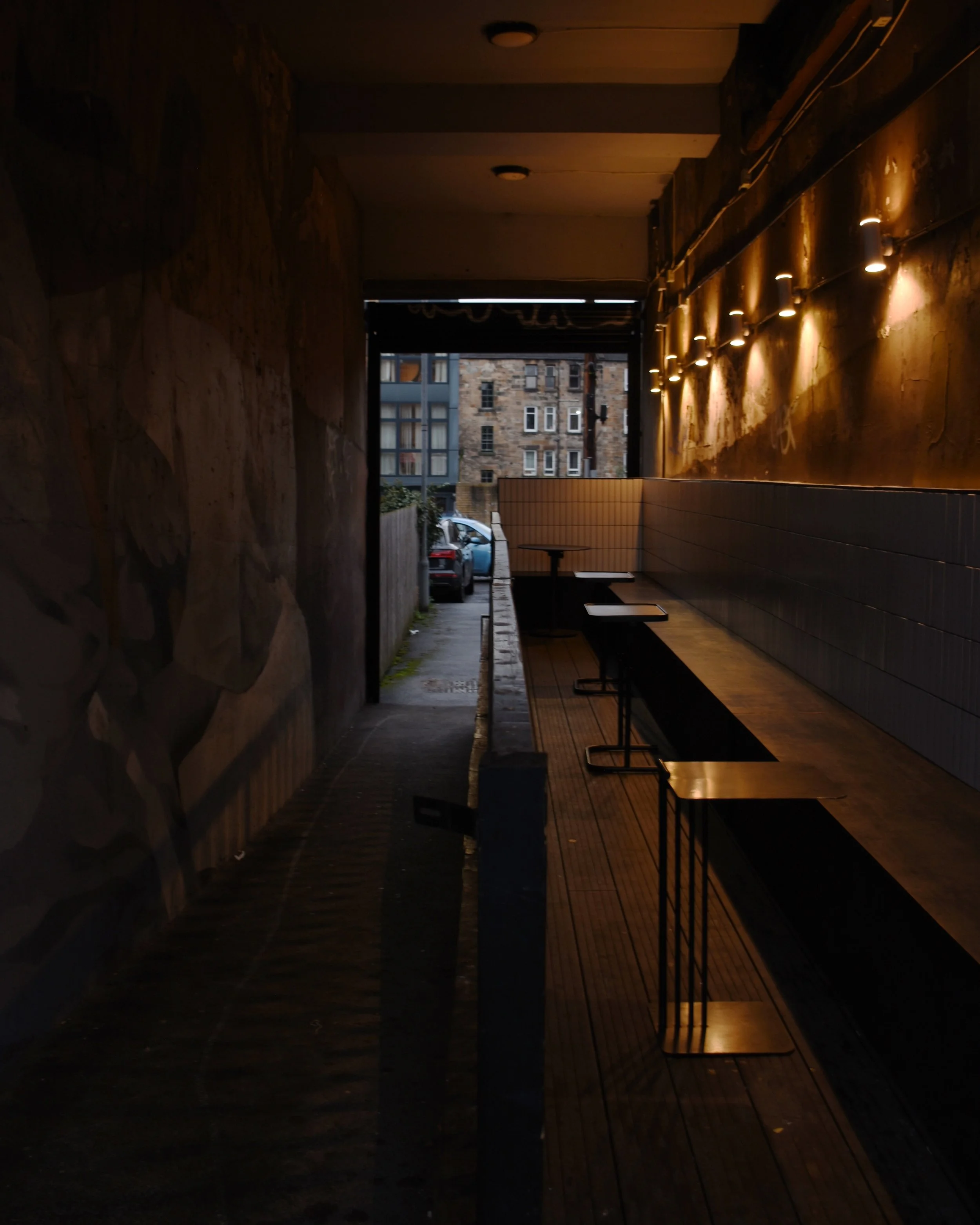 A dimly lit café interior with wooden tables and chairs, a textured wall with warm lighting, and an open door leading outside to a city street with parked cars and tall buildings.