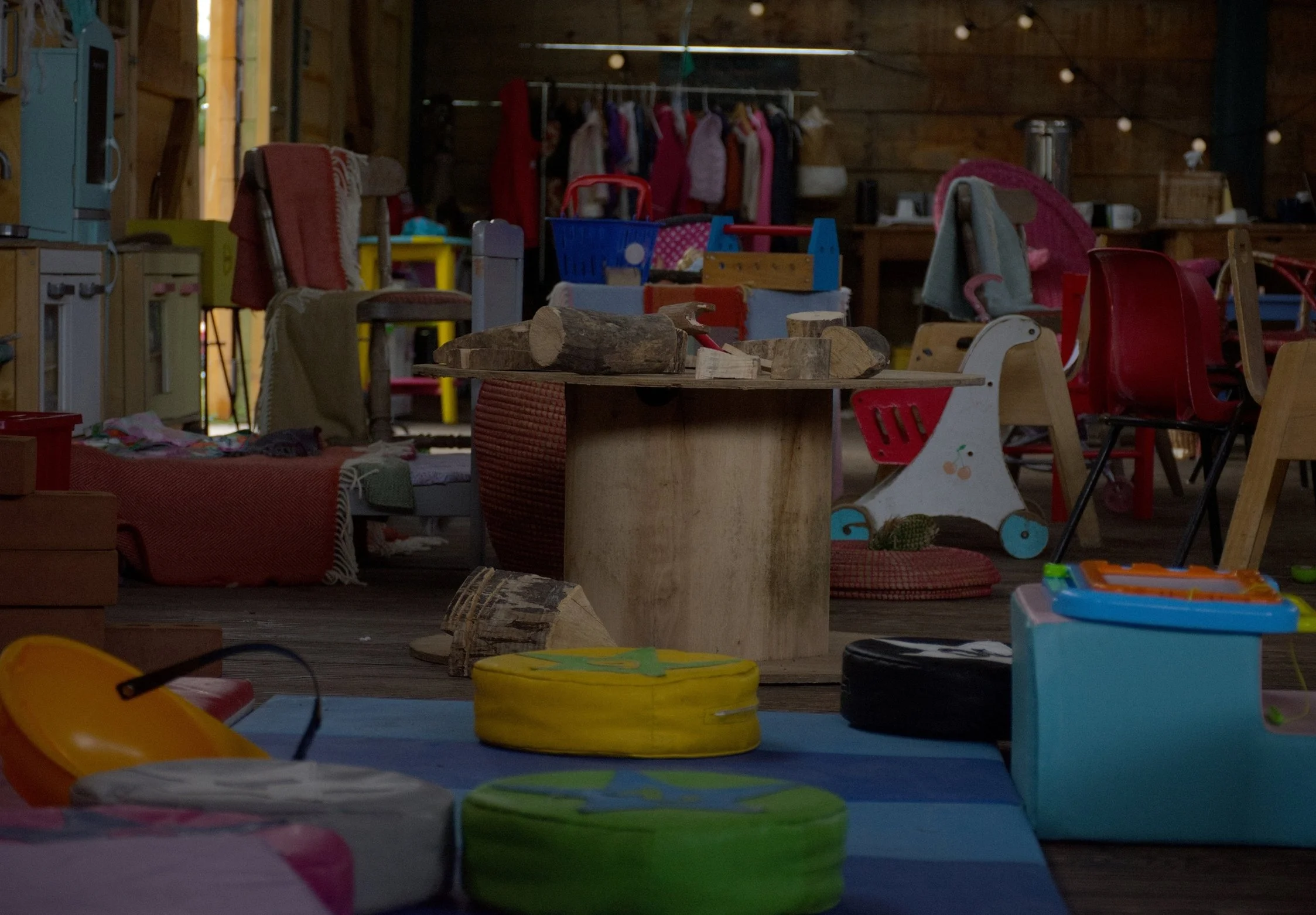 Children's toys scattered on a playroom floor with chairs, logs, and a wooden table in a rustic room decorated with hanging string lights and hanging clothes.