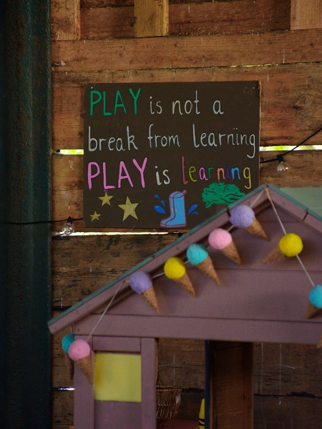 Decorative sign with the quote "Play is not a break from learning, play is learning" on a wooden wall, accompanied by illustrations of stars, a boot, and a tree. Part of a colorful stand with hanging faux ice cream cones is visible in the foreground.