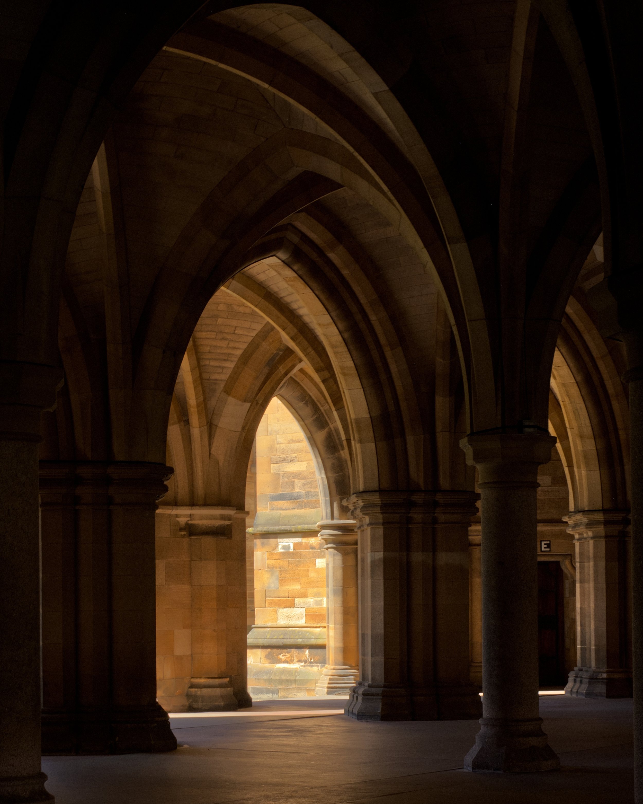 Interior University of Glasgow cloisters,  a historic stone building with arched ceilings and columns, illuminated by sunlight coming through an opening.