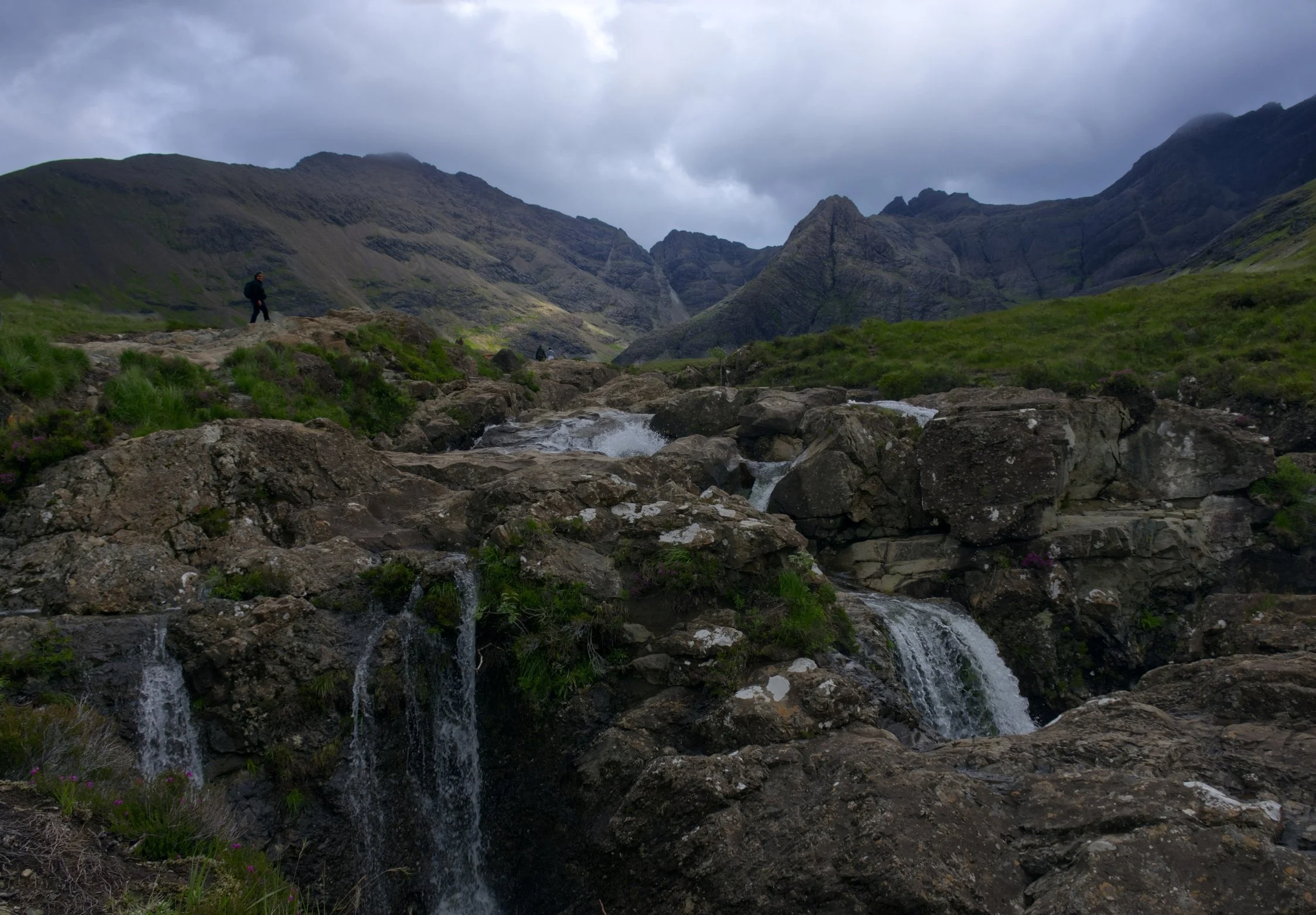 A hiker standing on rocky terrain near a small stream with waterfalls within a vast mountainous landscape under a cloudy sky.
