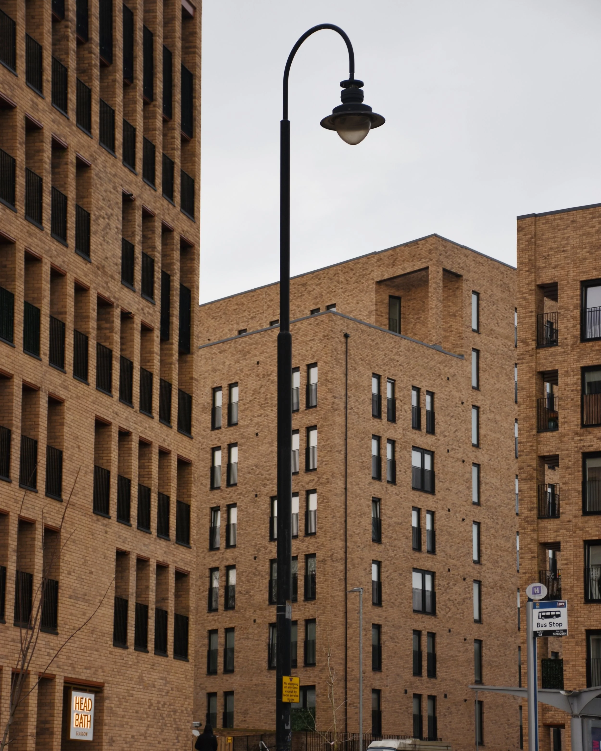 A street scene with a tall streetlight and multiple modern brown brick buildings with black window frames and small balconies. There are signs for a bus stop and a hotel. The sky is overcast.