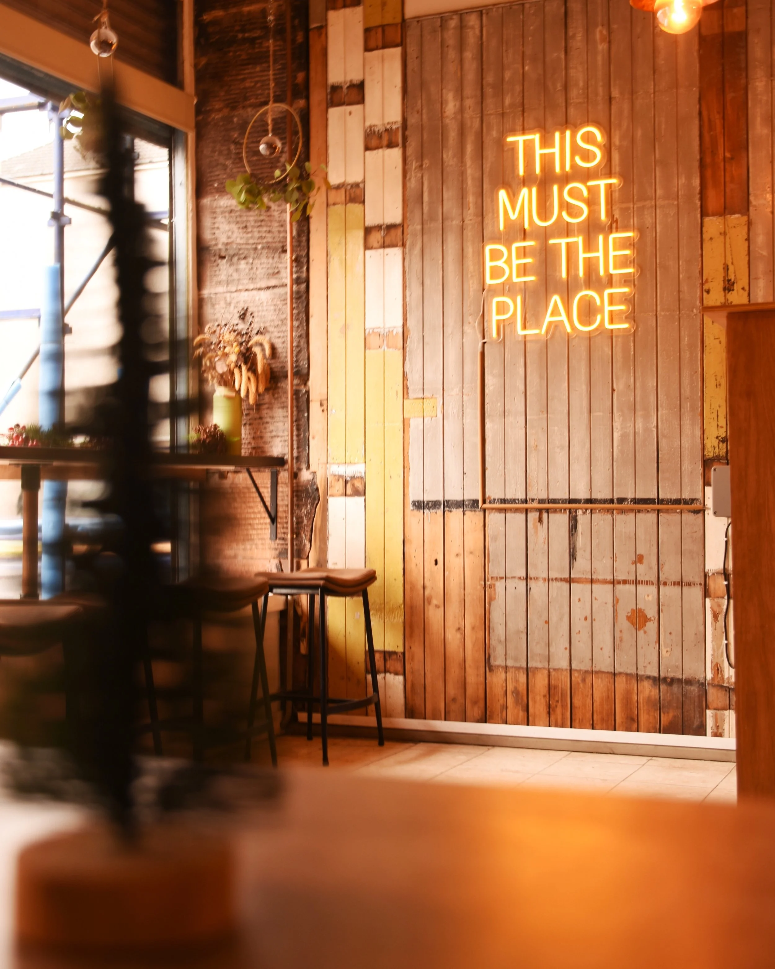 Interior of a cafe or restaurant with a wooden wall and a neon sign that reads 'This Must Be The Place'. There is a window on the left with plants on the windowsill, and some chairs and tables are partially visible.