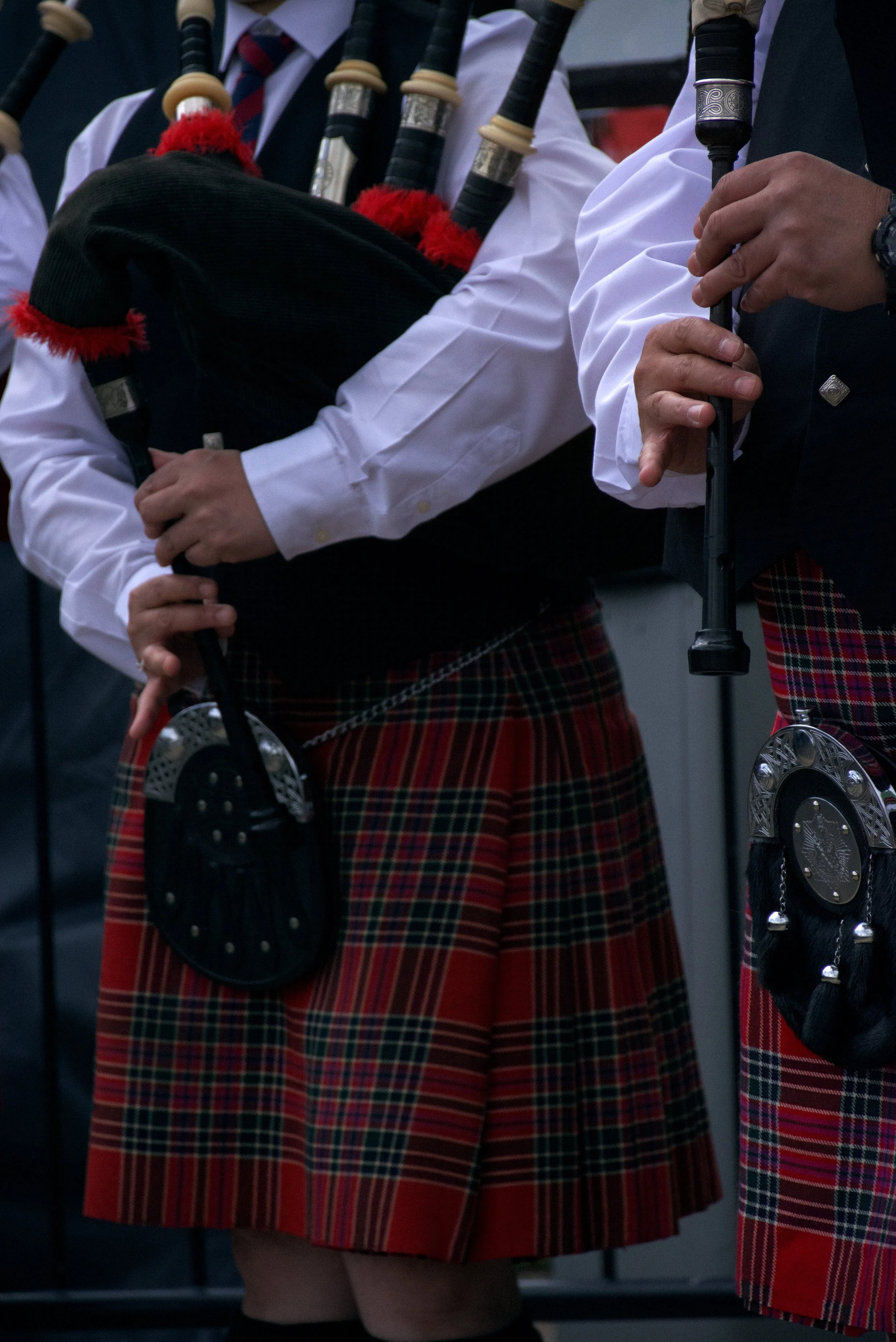 Close-up of musicians in traditional Scottish attire playing bagpipes, wearing kilt skirts with tartan patterns, white shirts, and holding bagpipes with black and silver accents.