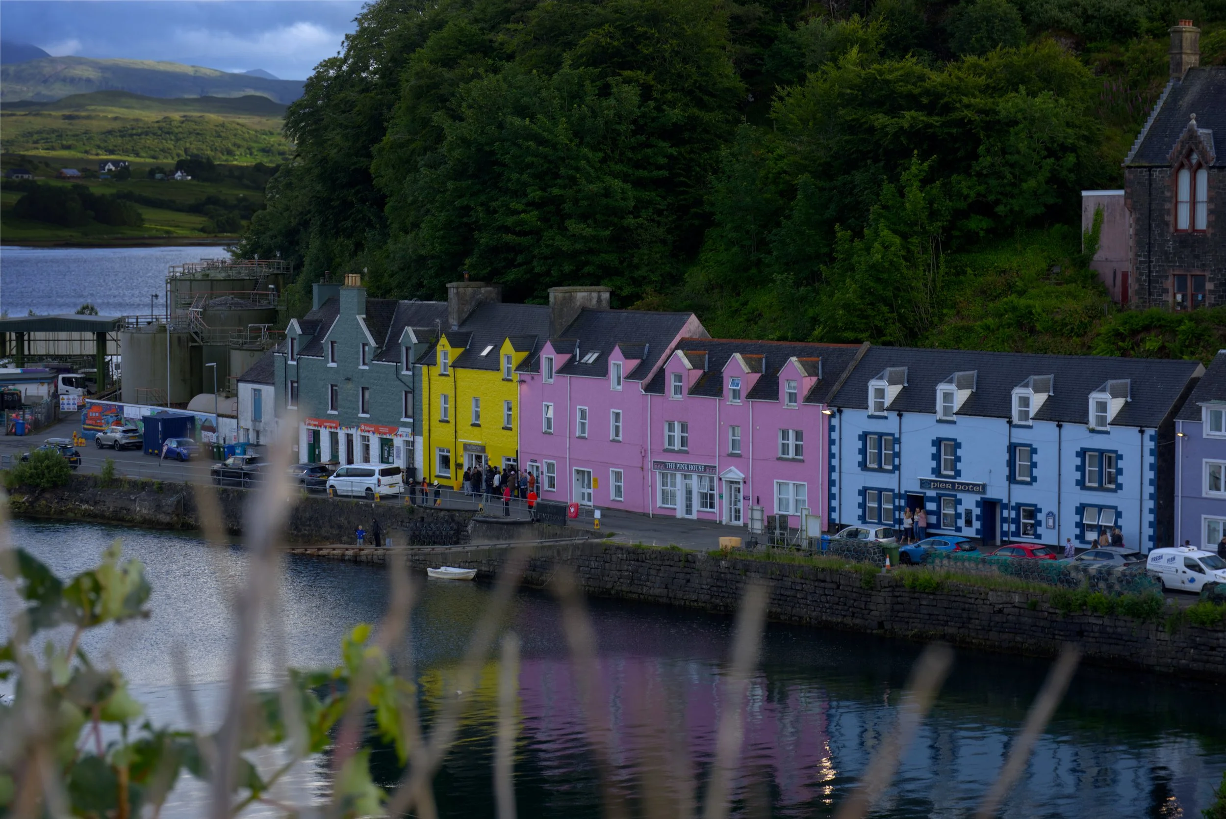 Portree on Skye, colourful buildings along the waterfront with boat and parked cars, set against green hills and trees under cloudy sky.