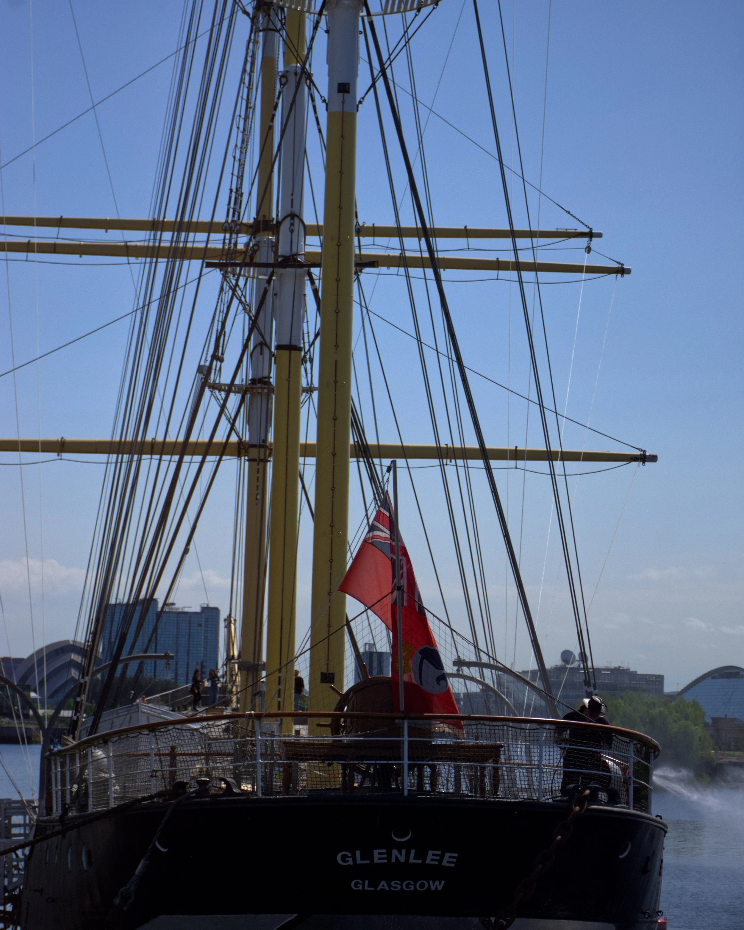 The Tall Ship Glasgow, a ship with multiple masts and rigging, city buildings and a modern architectural structure in the background. The sky is clear and blue.