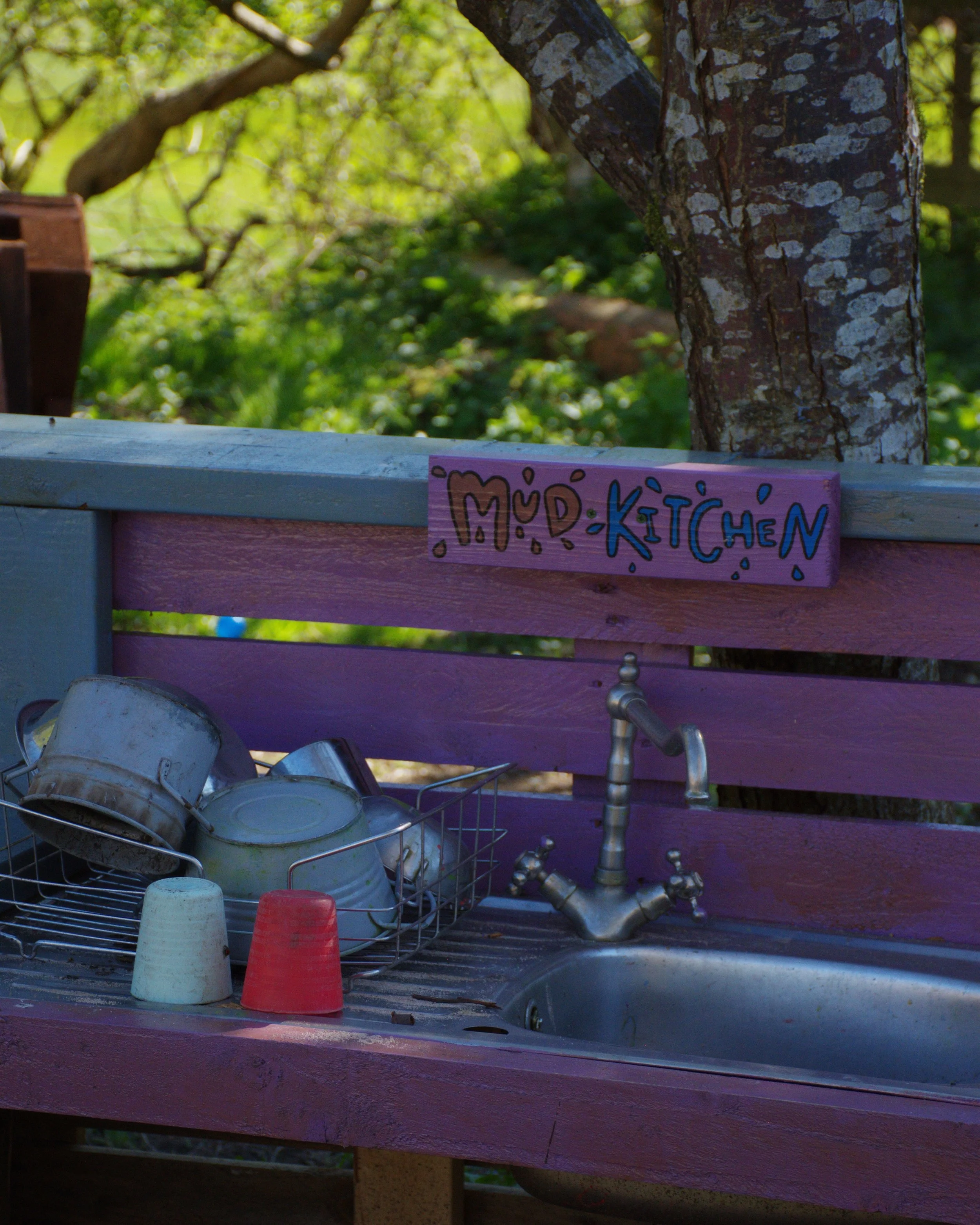 Outdoor kitchen area with a pink wooden sign that says 'MUD KITCHEN,' plastic cups on a rack, and a metal sink with a faucet, set against a blurred background of trees and greenery.
