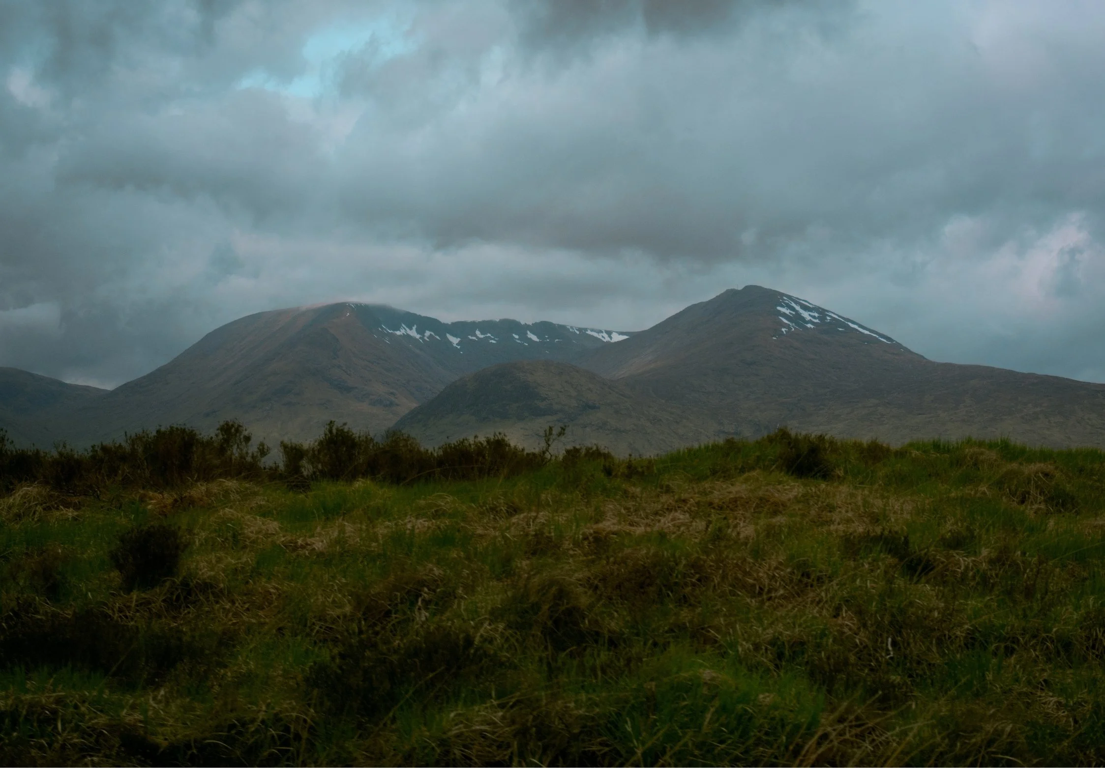 Glencoe landscape view of green grassy field with mountains in the background under a dark, cloudy sky.