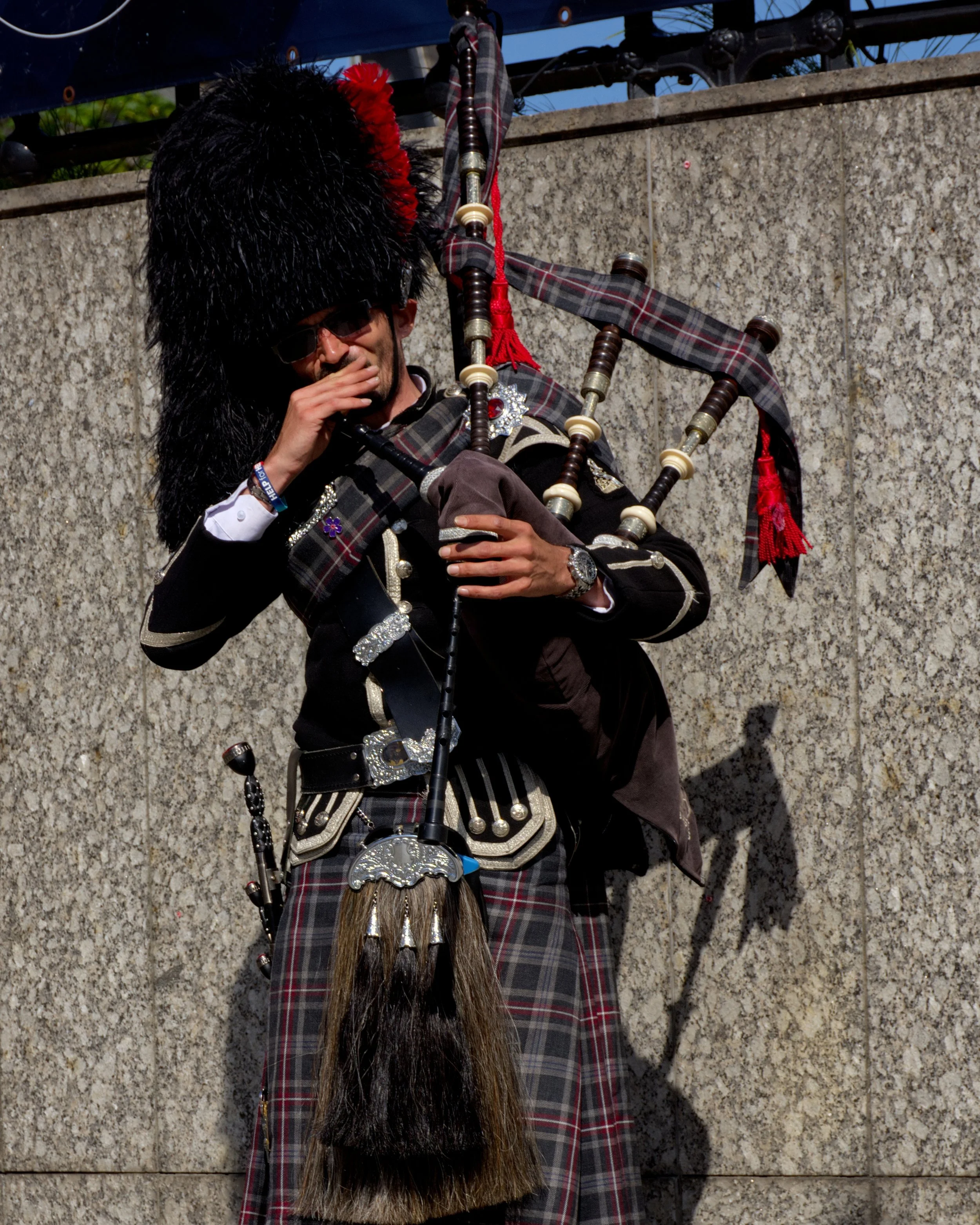 A man dressed in traditional Scottish attire playing bagpipes outdoors, wearing a black hat with a large feather, kilt, and jacket, with a stone wall in the background.