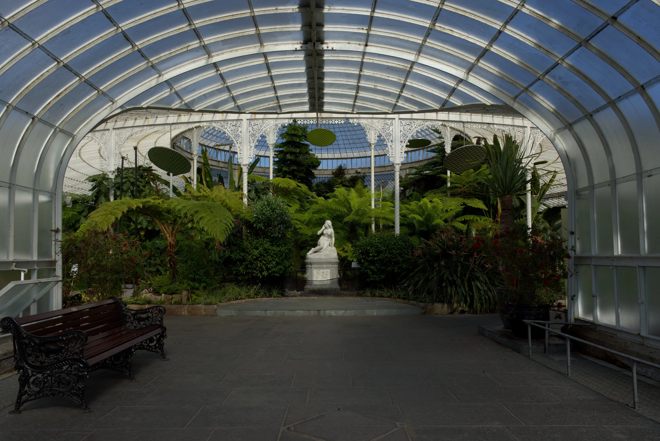 Interior of a the Glasgow botanical gardens, a glass conservatory filled with lush green plants and a white sculpture of a seated woman at the centre.