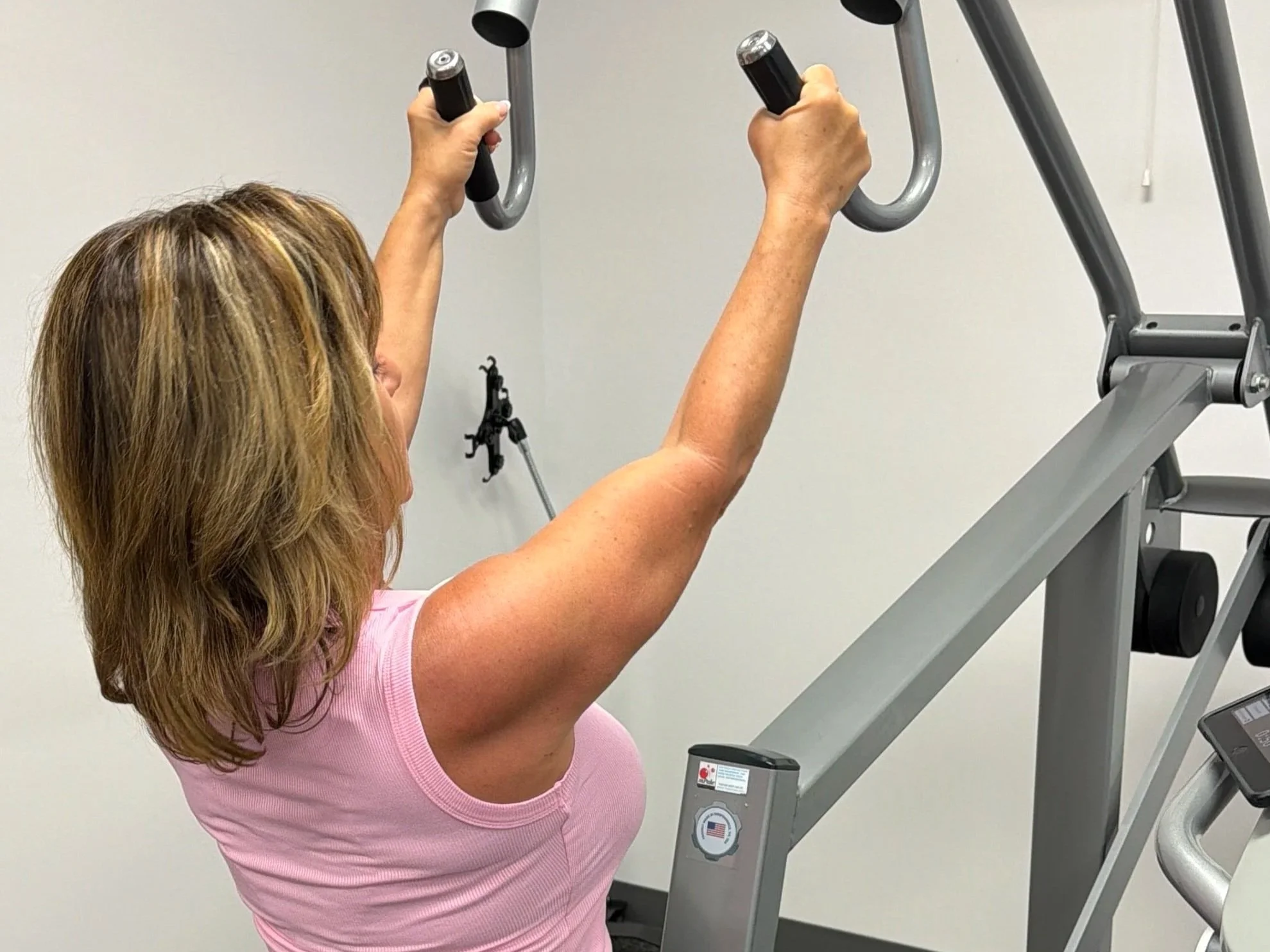 A woman in a pink shirt exercising using a piece of gym equipment with handlebars in an indoor fitness facility.