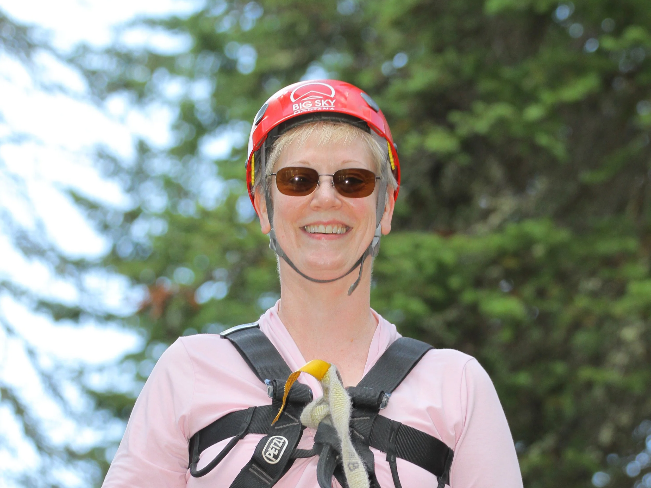 Woman wearing a red helmet with 'Big Sky' logo, sunglasses, and a pink shirt, smiling outdoors in a wooded area.
