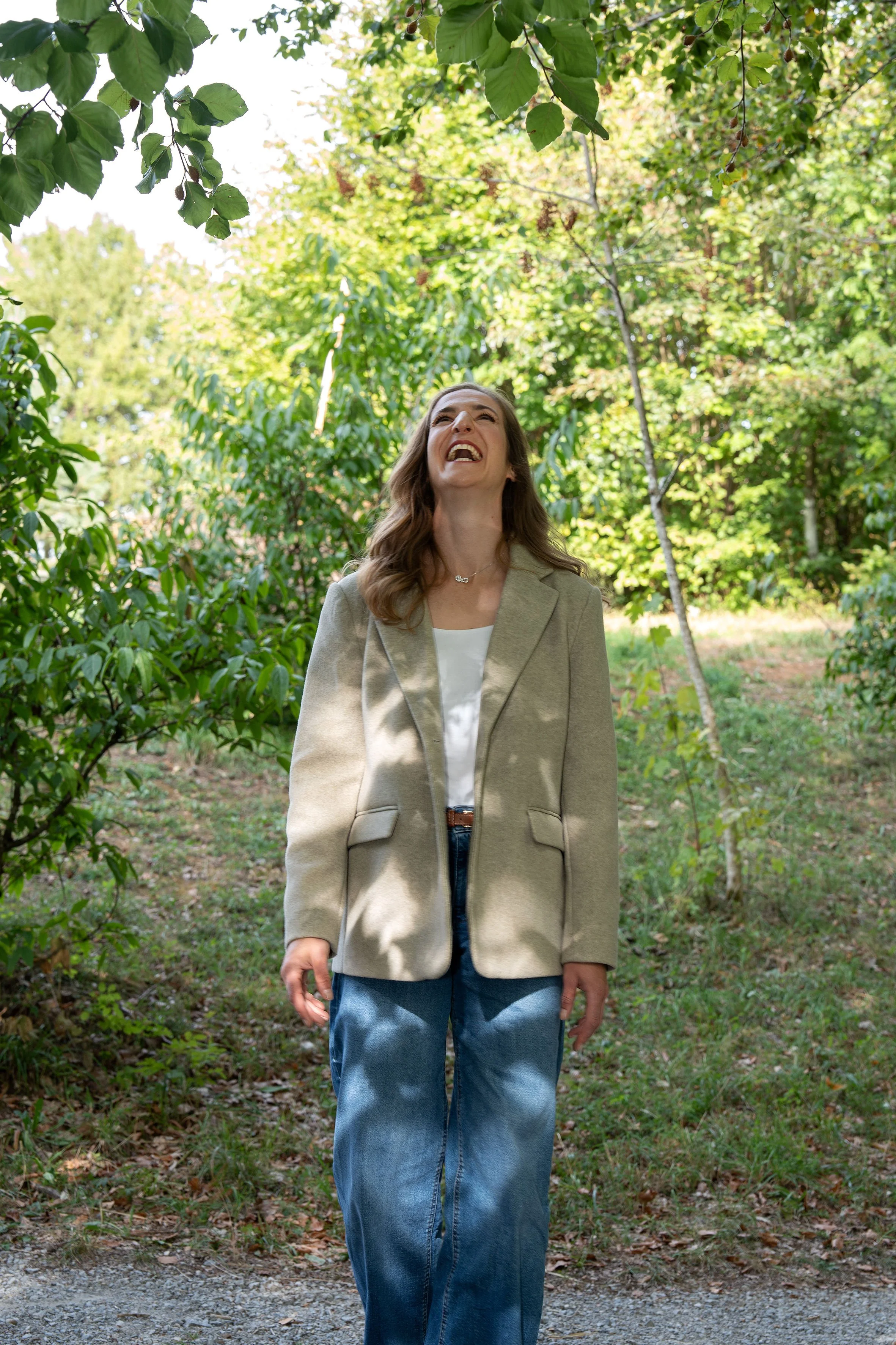 Eine Frau steht in einem grünen Wald und schaut nach oben, lacht und hat die Arme locker an den Seiten. Sie trägt eine beige Jacke und Jeans.