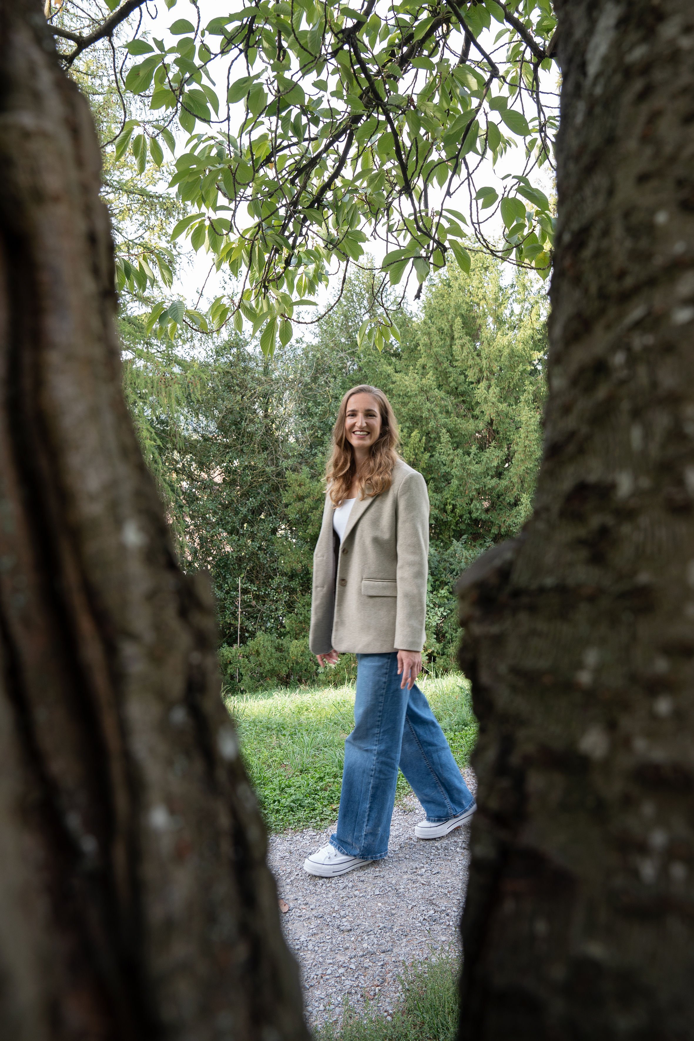 Eine lachende junge Frau mit langem braunem Haar, die in einem Park steht, gesehen durch eine Lücke zwischen zwei Baumstämmen.