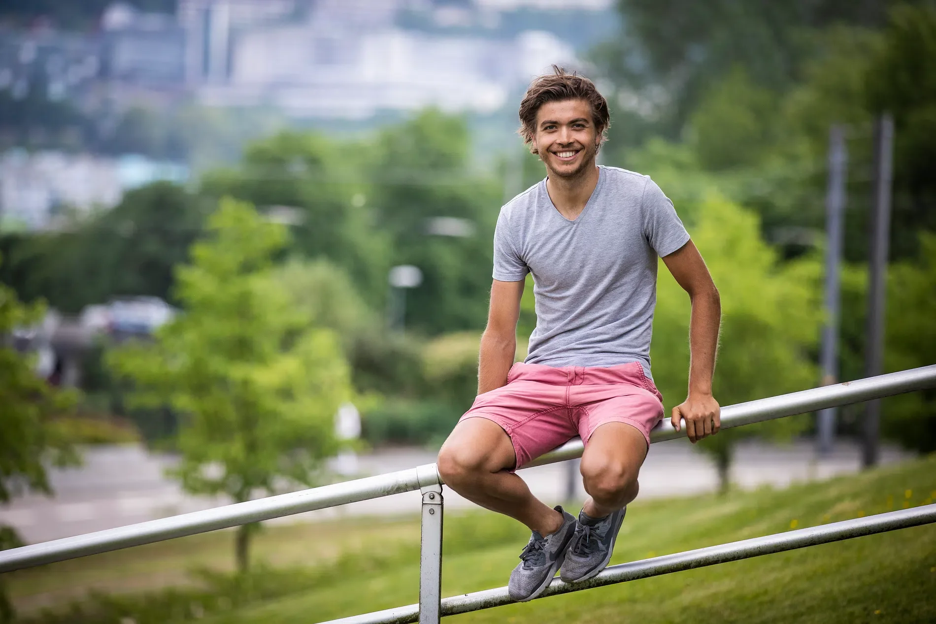 En smilende mann med skulderlangt hår, kledd i lysegrå t-skjorte og rosa shorts, sitter på en metallrekkverk i en grønn park eller urbane omgivelser.