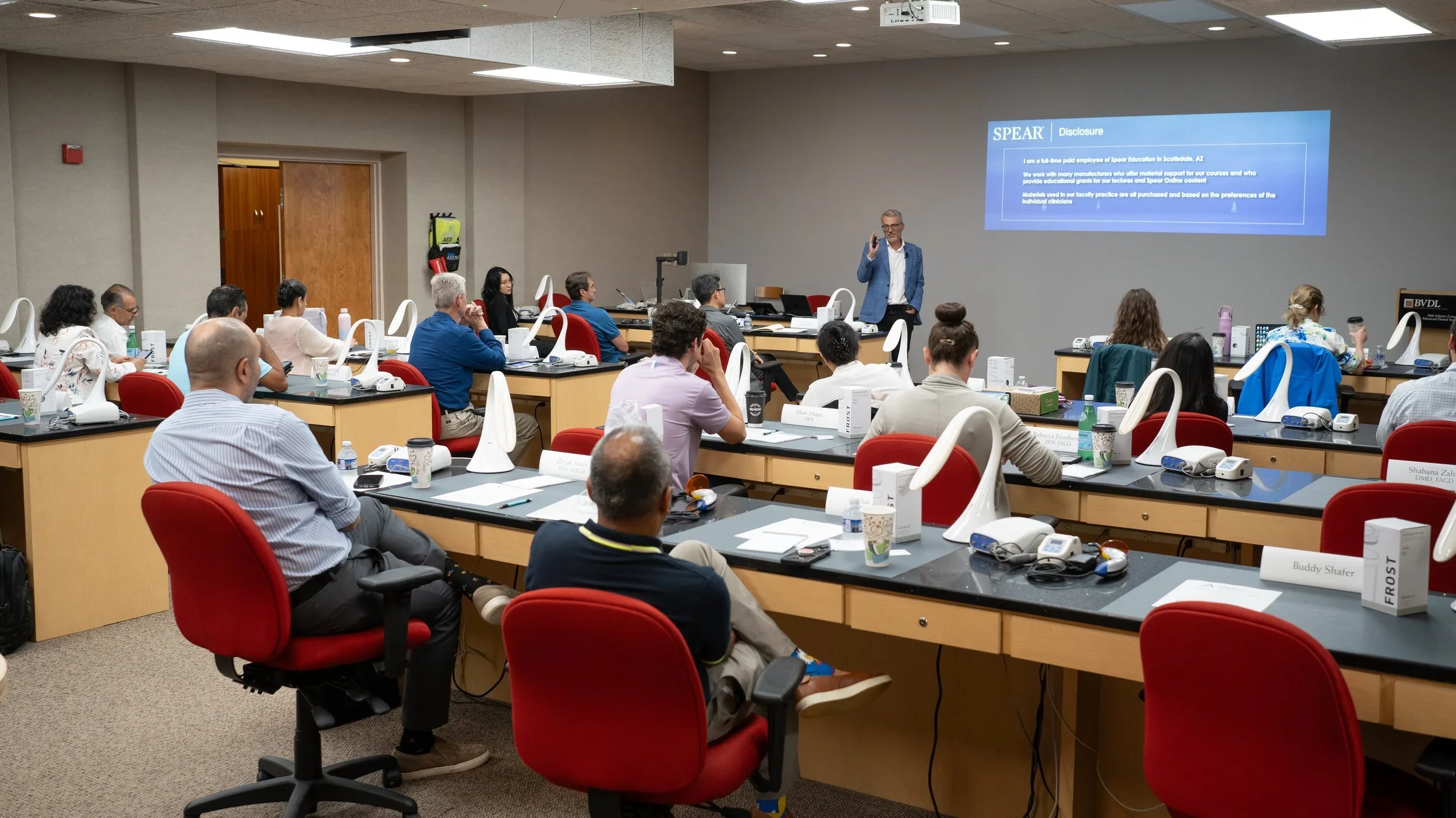 A conference room with attendees seated at desks, watching a speaker at the front, with a large screen displaying a slide titled 'SPEAR Disclosure'.