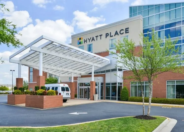 Exterior of a Hyatt Place hotel with a covered entrance, a small white shuttle bus, and landscaped greenery.