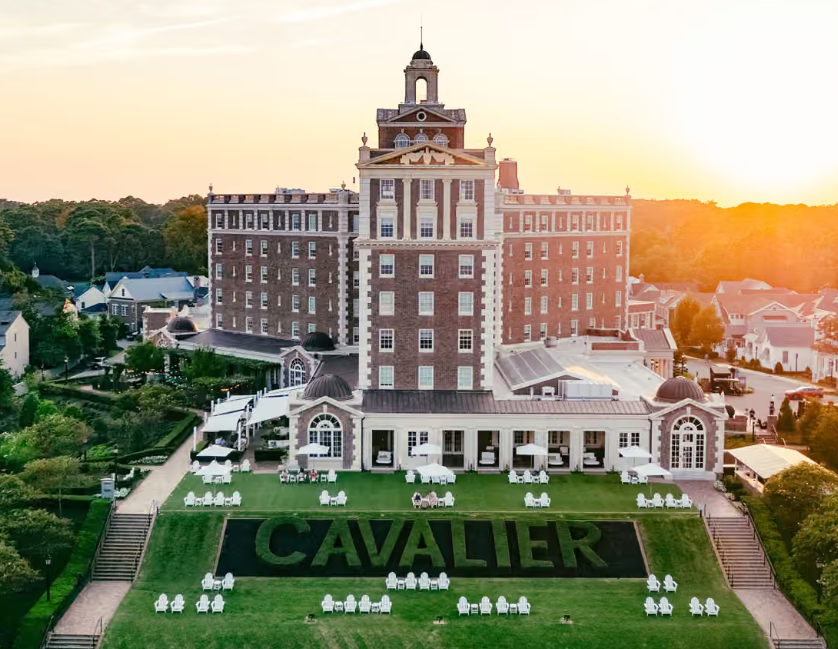 A large, historic hotel building with a prominent clock tower at sunset, situated on a landscaped hillside with the word "CAVALIER" spelled out in greenery on the lawn.