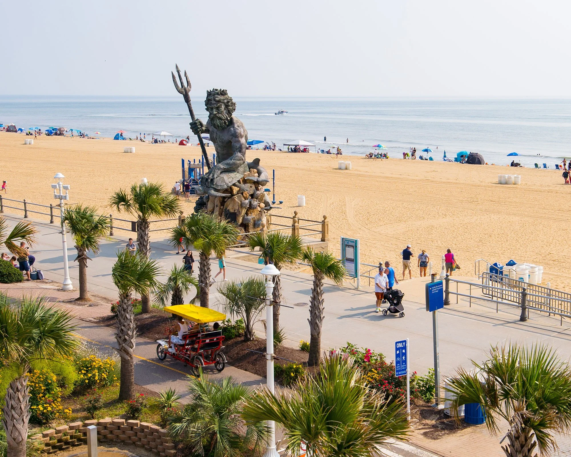 Beach with a large statue of Poseidon holding a trident in front, people on the sandy shore, umbrellas and boats on the ocean, and a walkway with palm trees and a pedal-powered trolley with passengers.