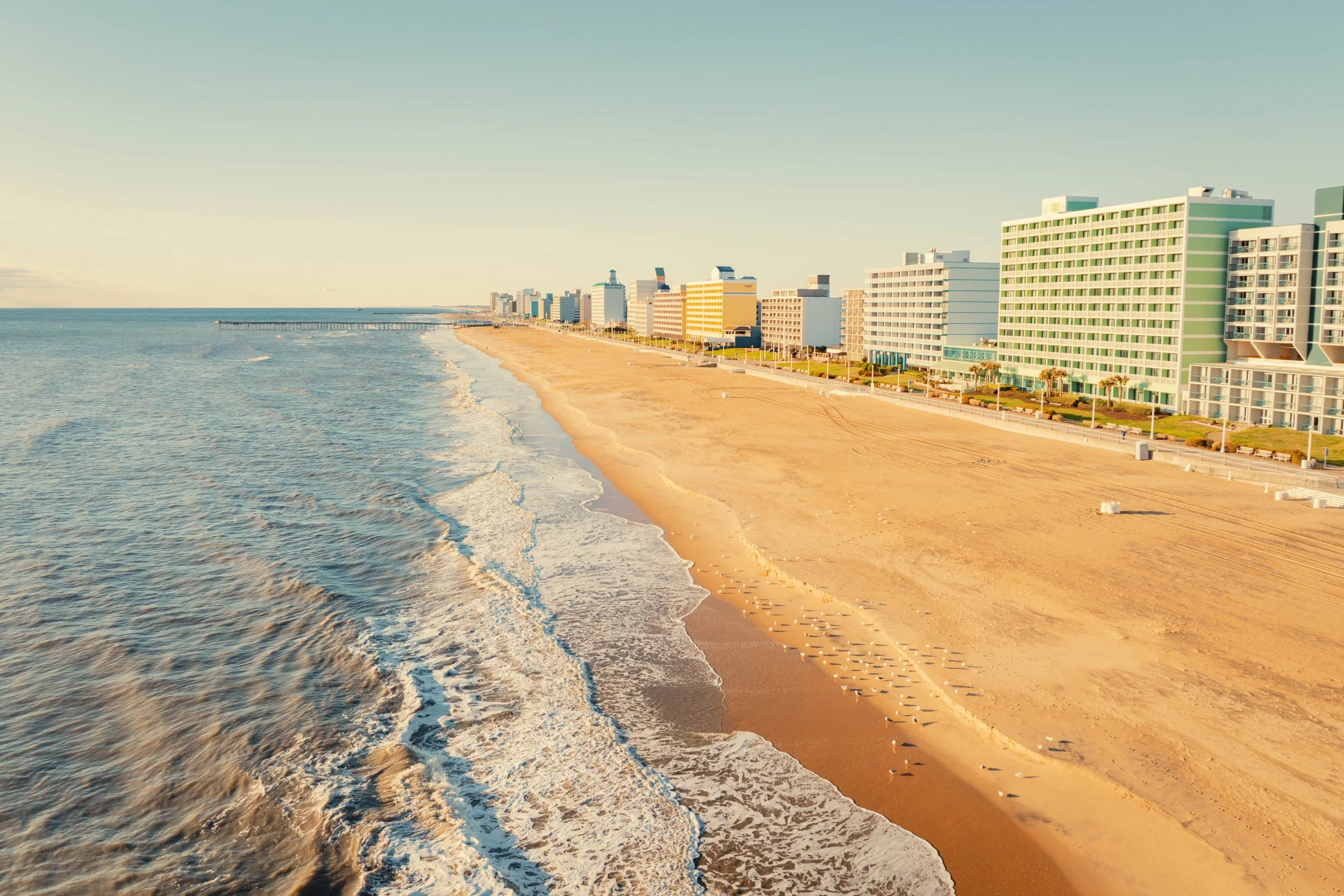 A sandy beach with gentle waves washing ashore, lined with tall modern hotels and condos under a clear sky.