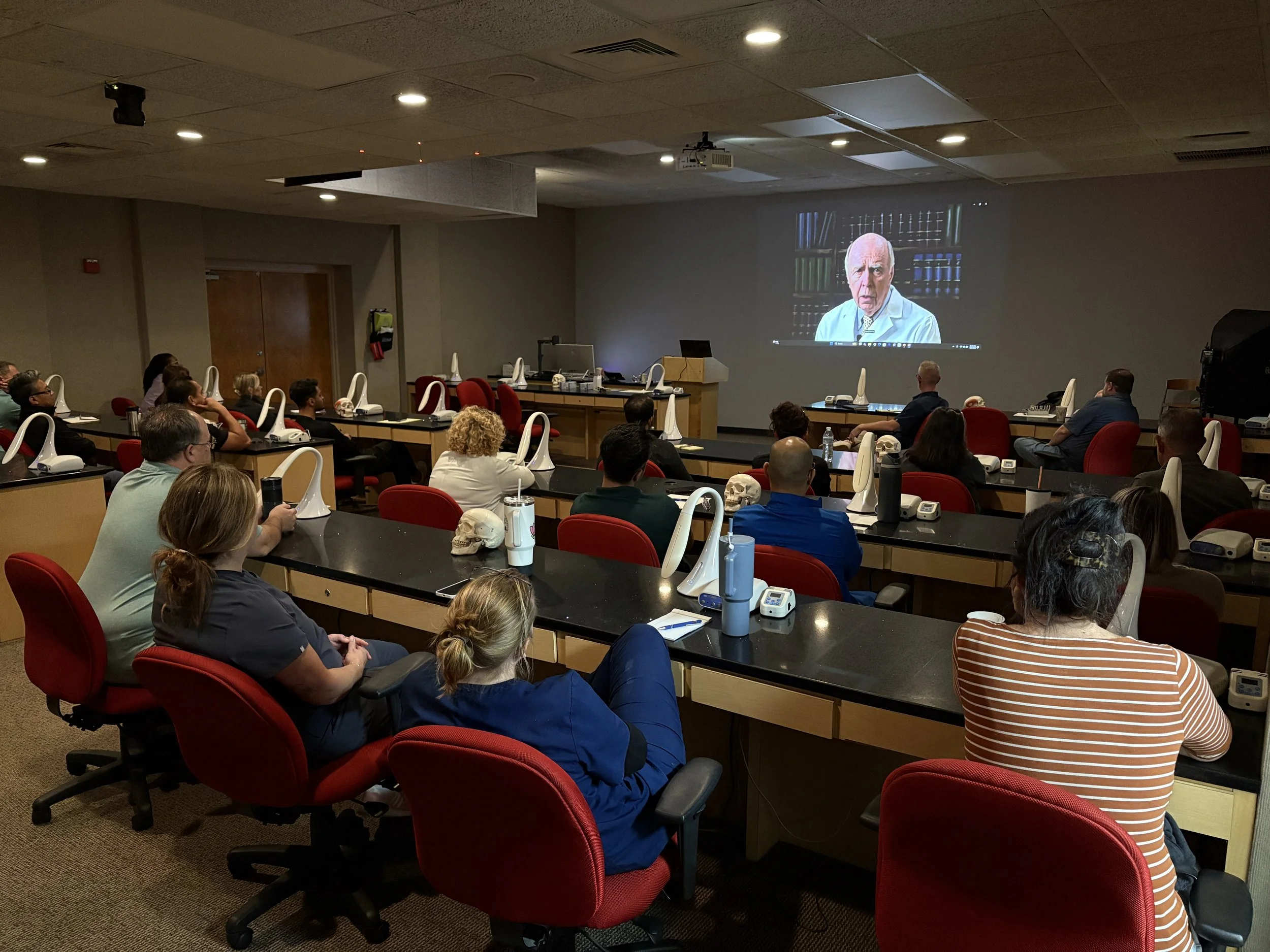 A group of people sitting in a conference room watching a presentation on a large screen. The screen shows an older man in a lab coat with bookshelves in the background.