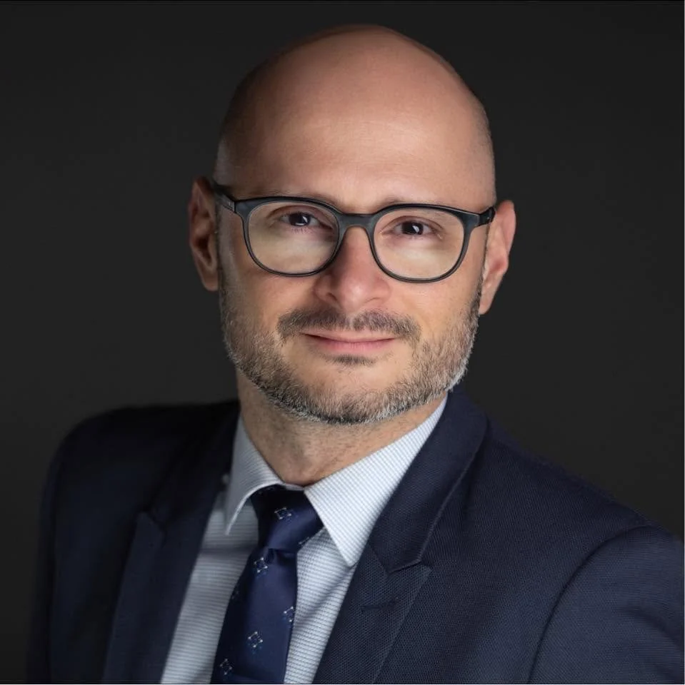 Portrait of a bald man with glasses, wearing a navy suit, light blue shirt, and dark tie, smiling against a dark background.