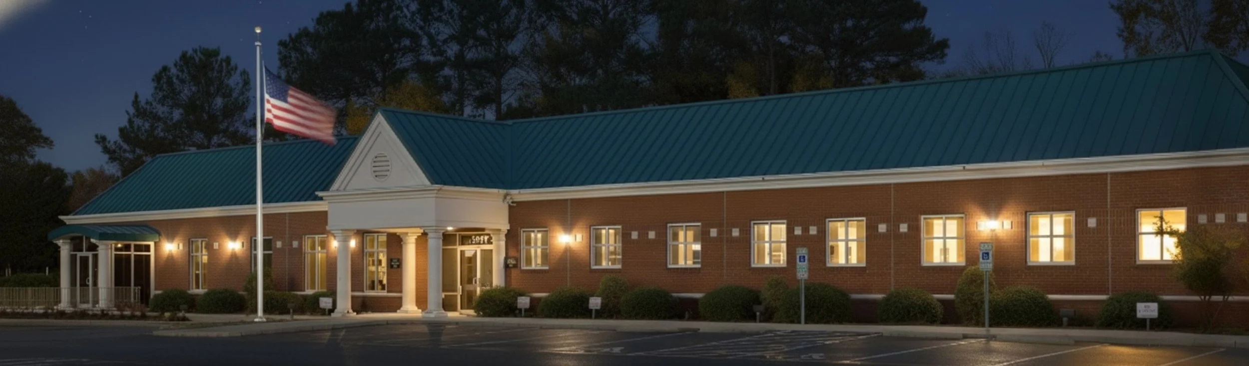 A brick building at night with illuminated windows and a flagpole with the American flag in front.