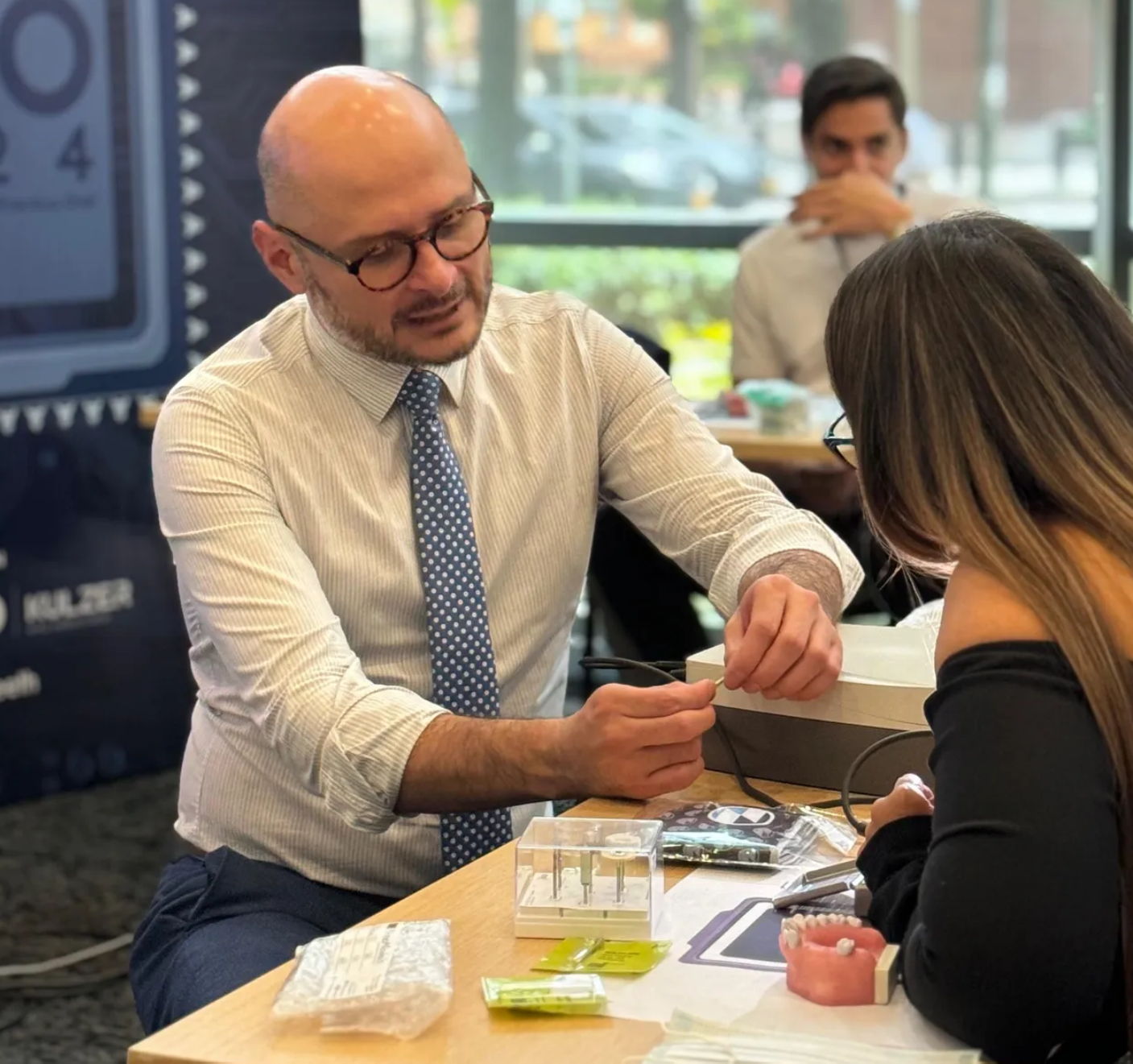 A man with glasses and a beard, dressed in a light shirt and polka dot tie, is demonstrating a dental tool on a woman with long hair and glasses. They are seated at a table with dental models and supplies, indoors with a window showing a parking lot outside.