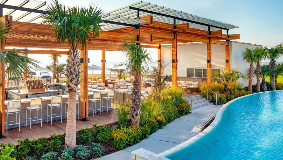 Poolside outdoor bar and dining area with palm trees, wooden structures, and a curved pool under a sunny sky.