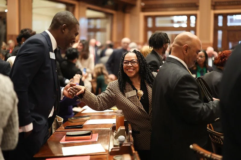 A woman shaking hands with a man at a professional event or conference; she is smiling and wearing glasses, and there are many people in the background.