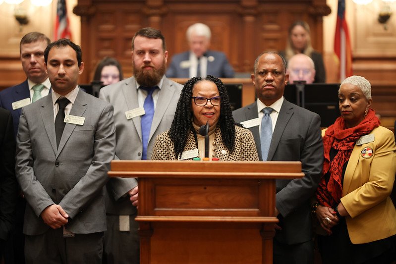 A woman speaking at a podium with microphones, surrounded by five other people, in a formal setting resembling a courtroom or official government chamber.