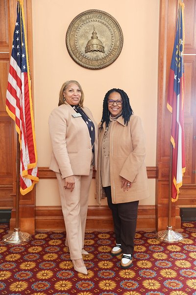 Two women standing side by side in front of a beige wall, with the Georgia state seal above them and flags of the United States and Georgia on either side.
