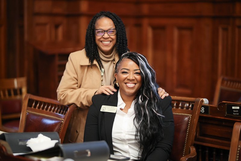 Two women in professional attire smiling in a wood-paneled room with desks and nameplates, one standing and the other sitting.