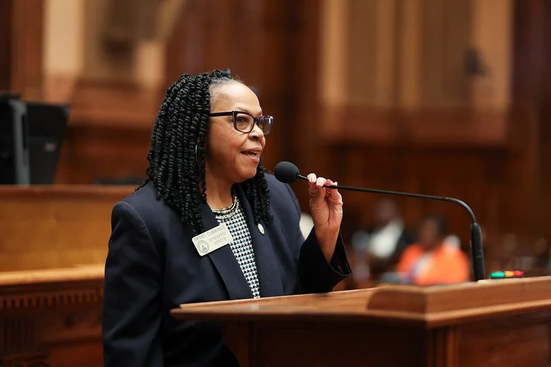 A woman with glasses, wearing a dark blazer and a patterned blouse, speaking into a microphone at a wooden podium in a courtroom or legislative chamber.
