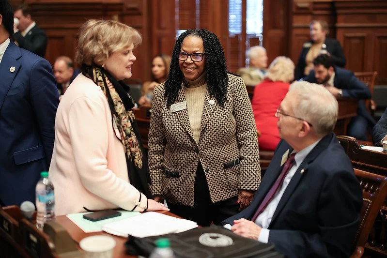 Three people are engaged in conversation inside a wood-paneled room filled with seated attendees; one woman is wearing a pink coat and scarf, another woman is wearing a patterned blazer with glasses, and a man is seated wearing a suit and glasses.
