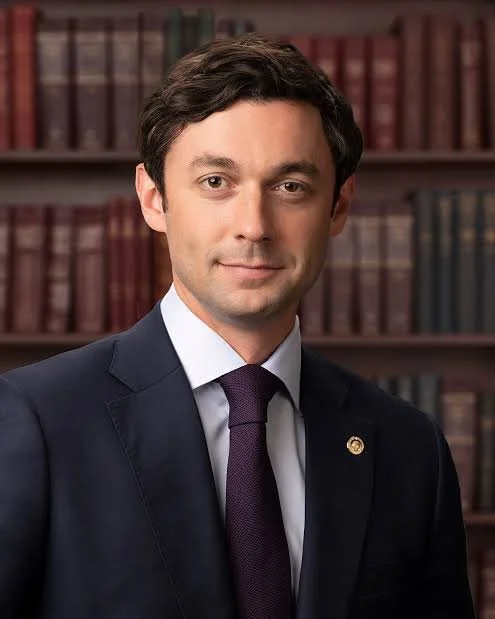 A man in a dark suit and tie standing in front of a background of bookshelves filled with books.