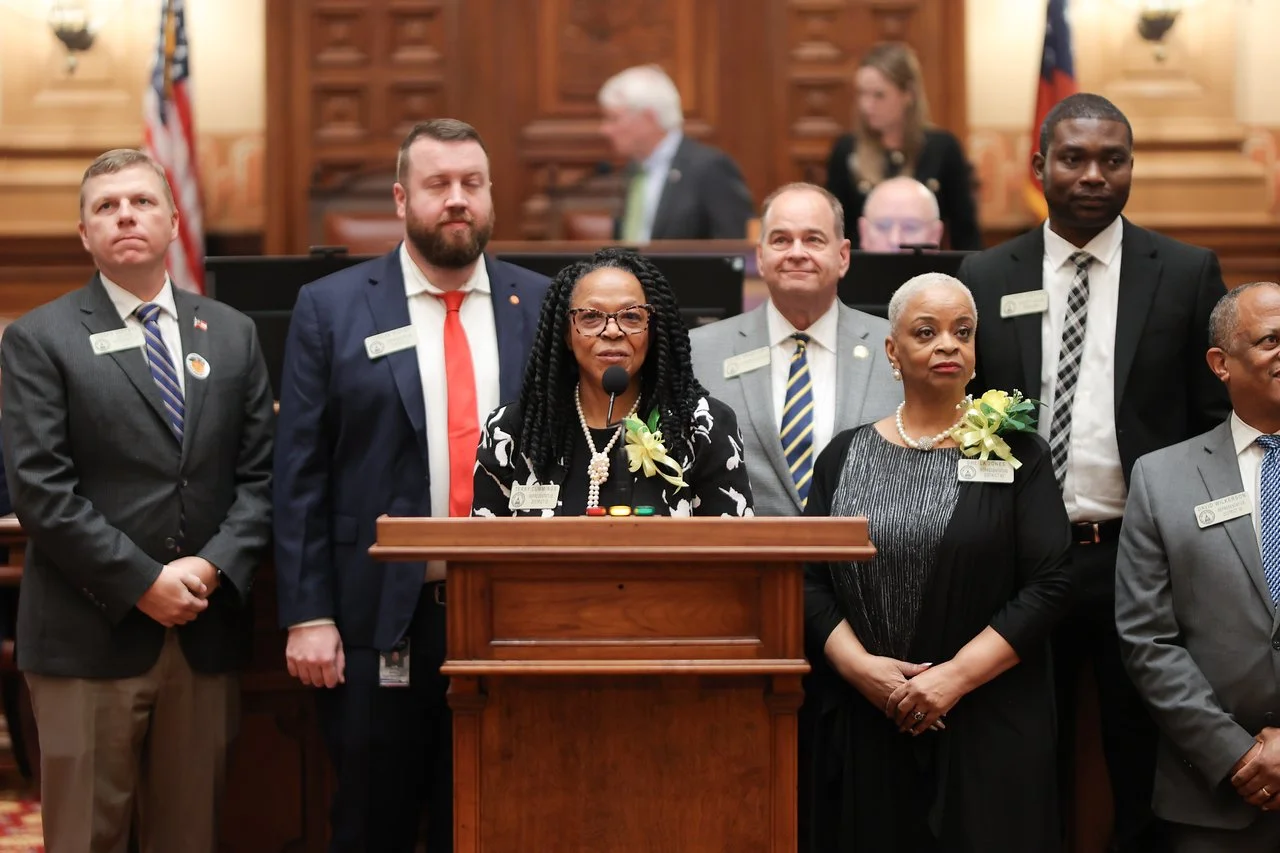 A group of diverse people standing behind a wooden podium during an official event in a formal setting, with some individuals wearing name tags and corsages.